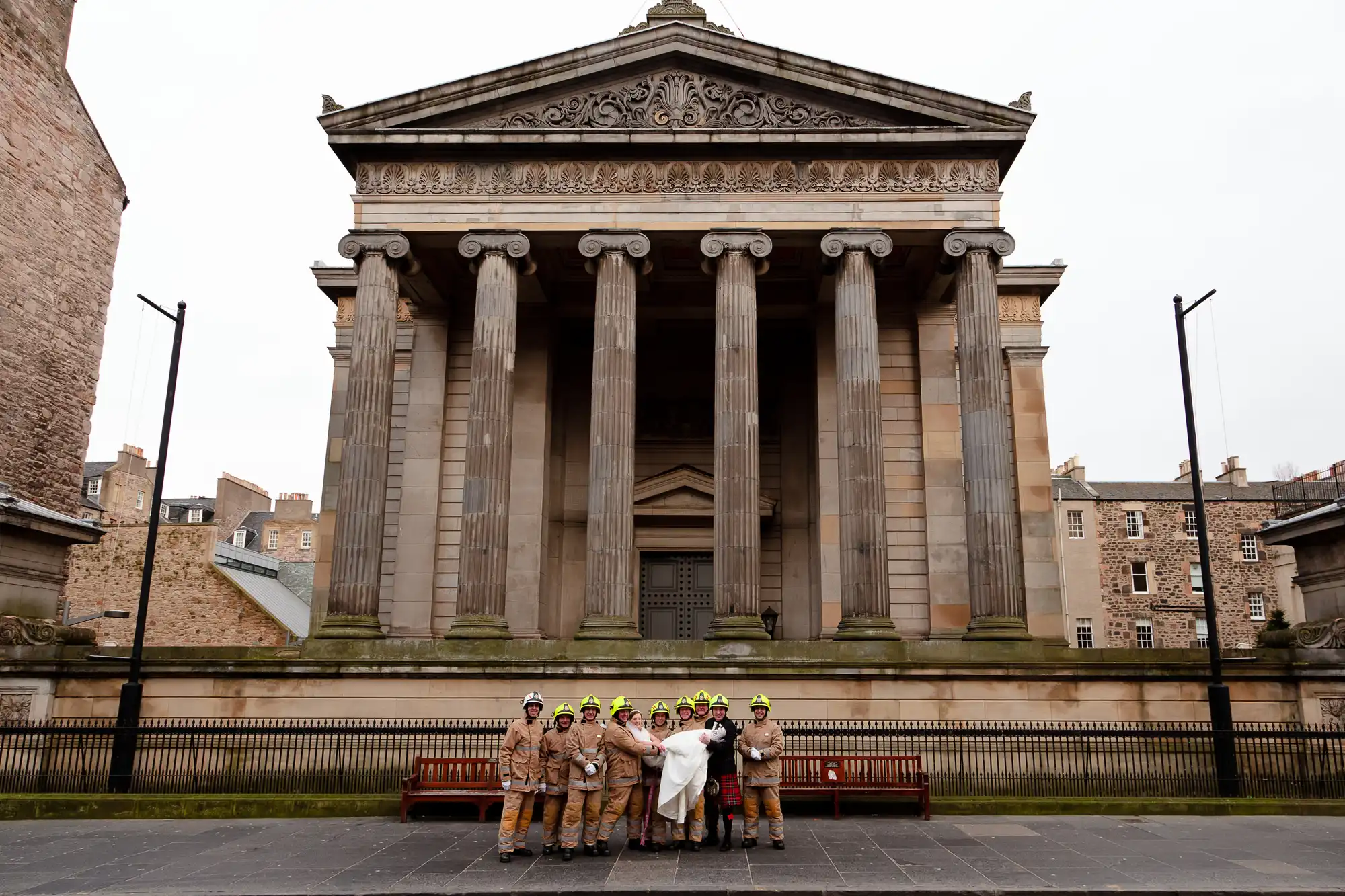A group of firefighters in full gear posing in front of an imposing neoclassical building with a large triangular pediment and columns.