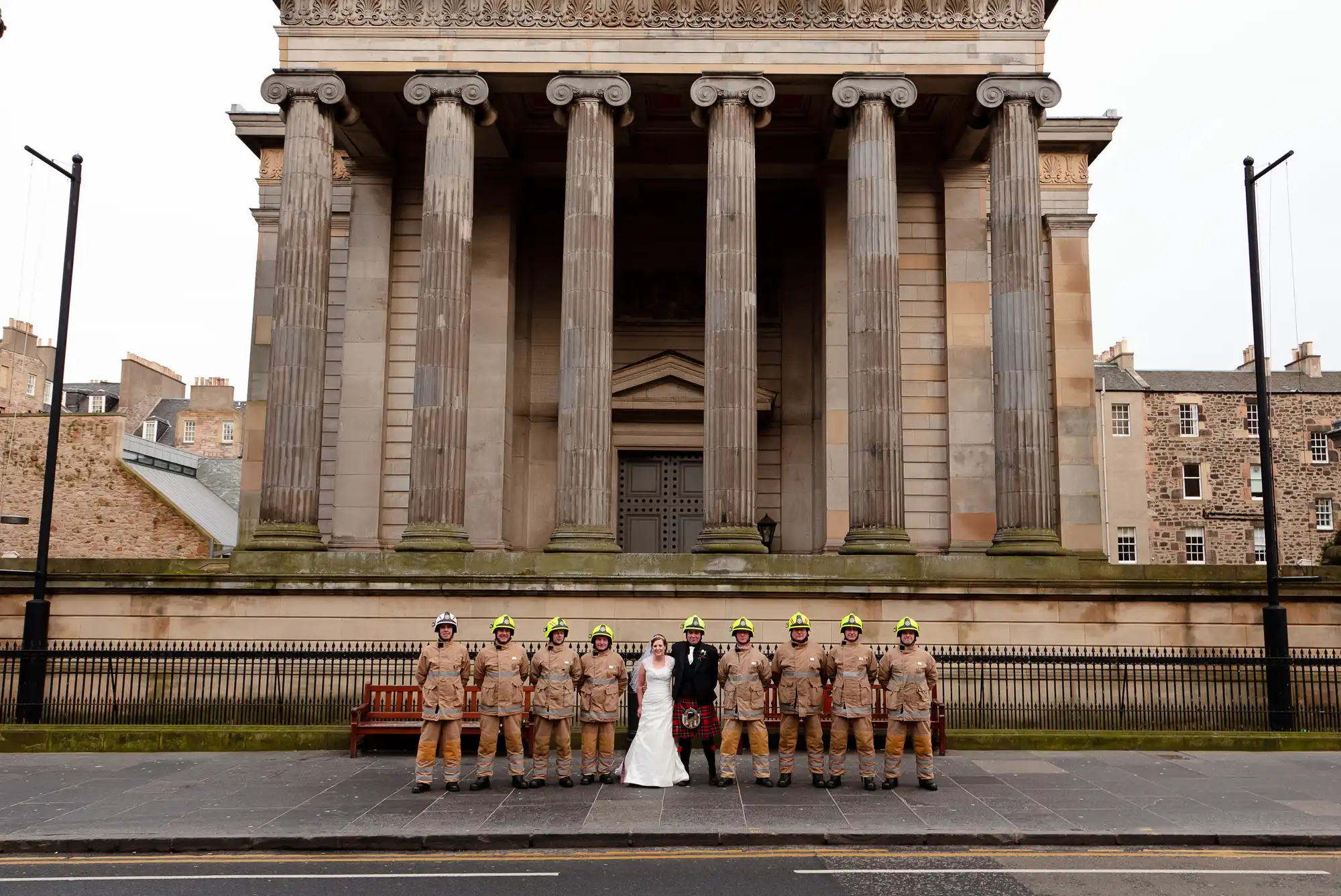 A bride and groom pose with a group of firefighters in front of the grand entrance of a classical stone building with columns.