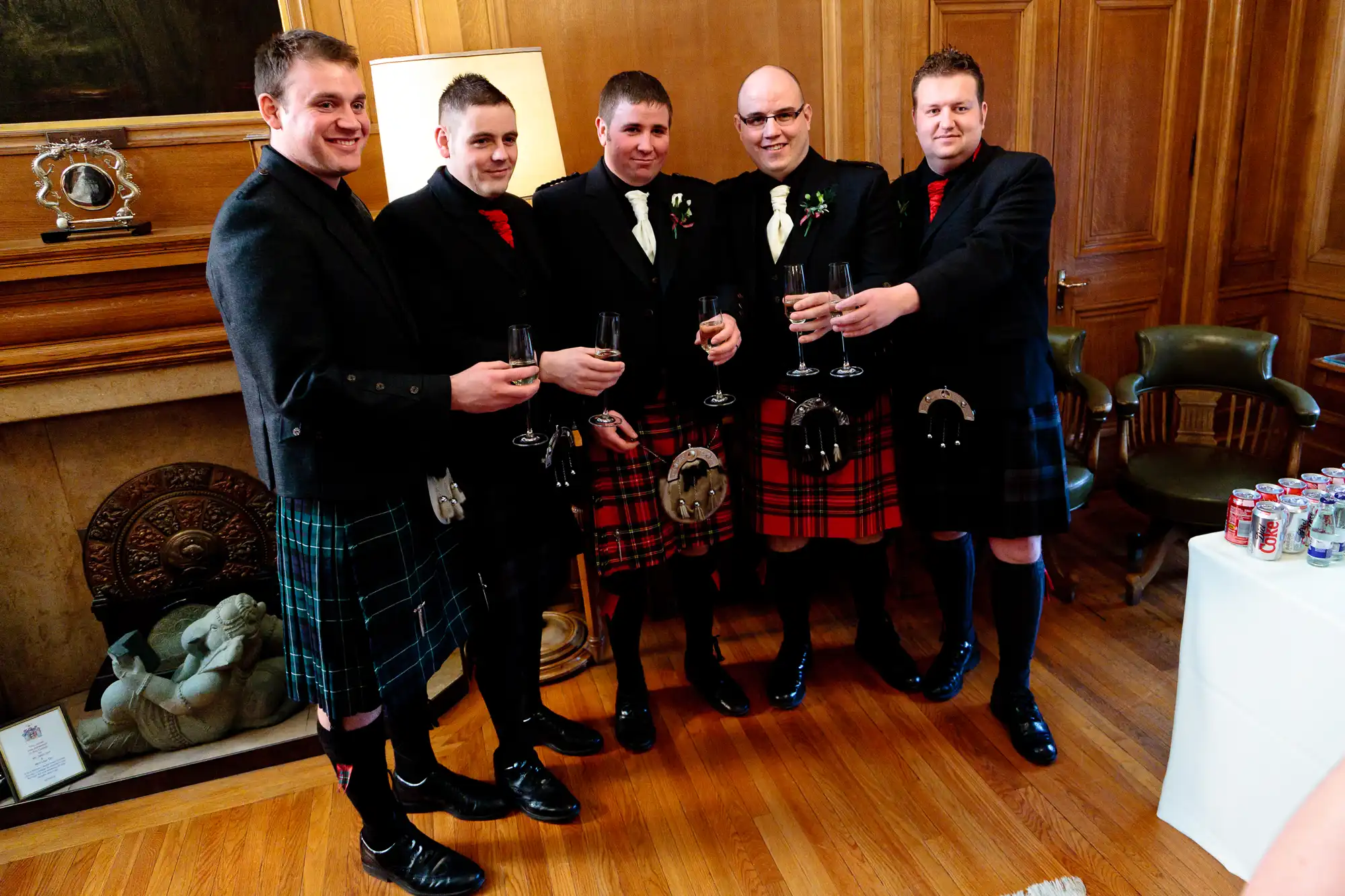 Five men in traditional scottish kilts and jackets, holding champagne glasses and smiling at a formal indoor event.