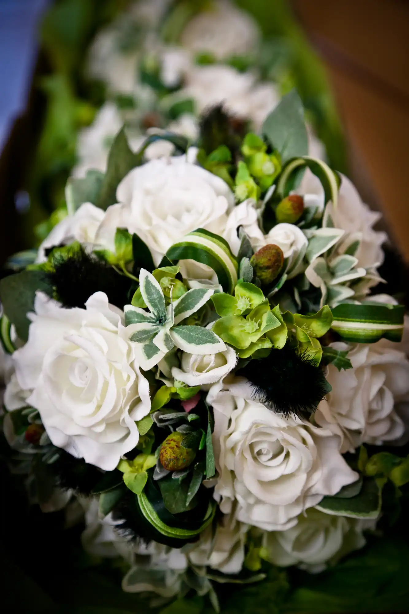 Close-up of a bouquet featuring white roses, green leaves, and fuzzy black details, with a blurred background.