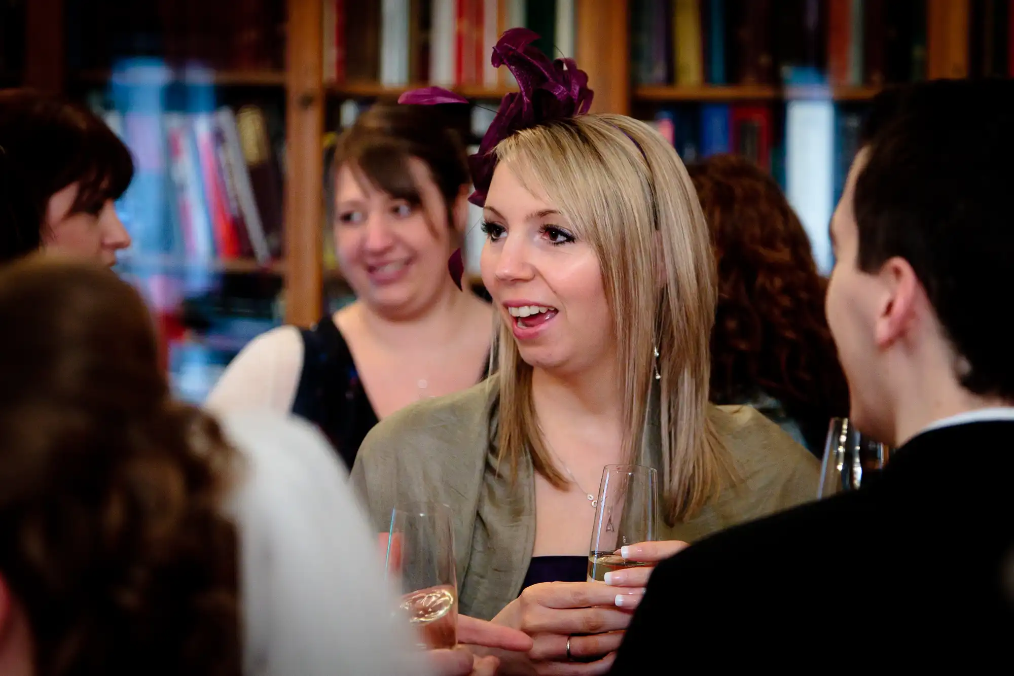 A woman with a purple fascinator talks animatedly with a group of people at a social gathering inside a room filled with bookshelves.
