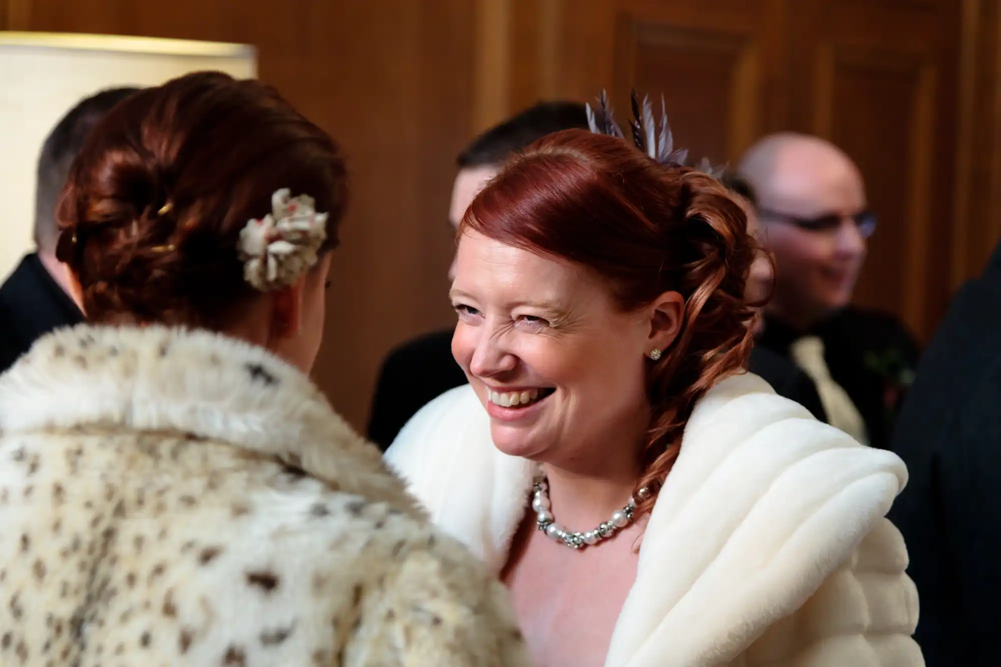 Two women sharing a joyful moment at a formal event, one wearing a white fur wrap and both with elaborate hairstyles.
