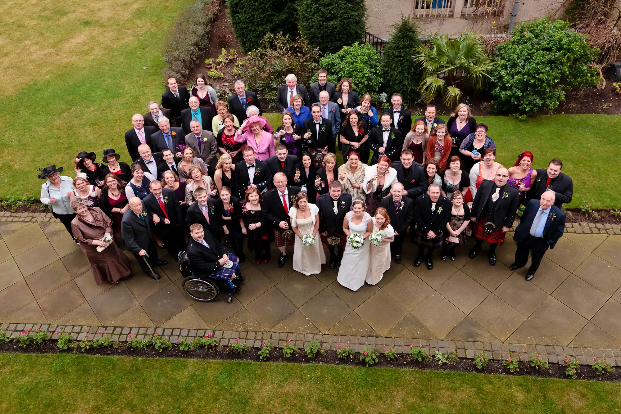 An aerial view of a large wedding group posing in a garden, featuring a bride and groom in the center surrounded by guests in formal attire.