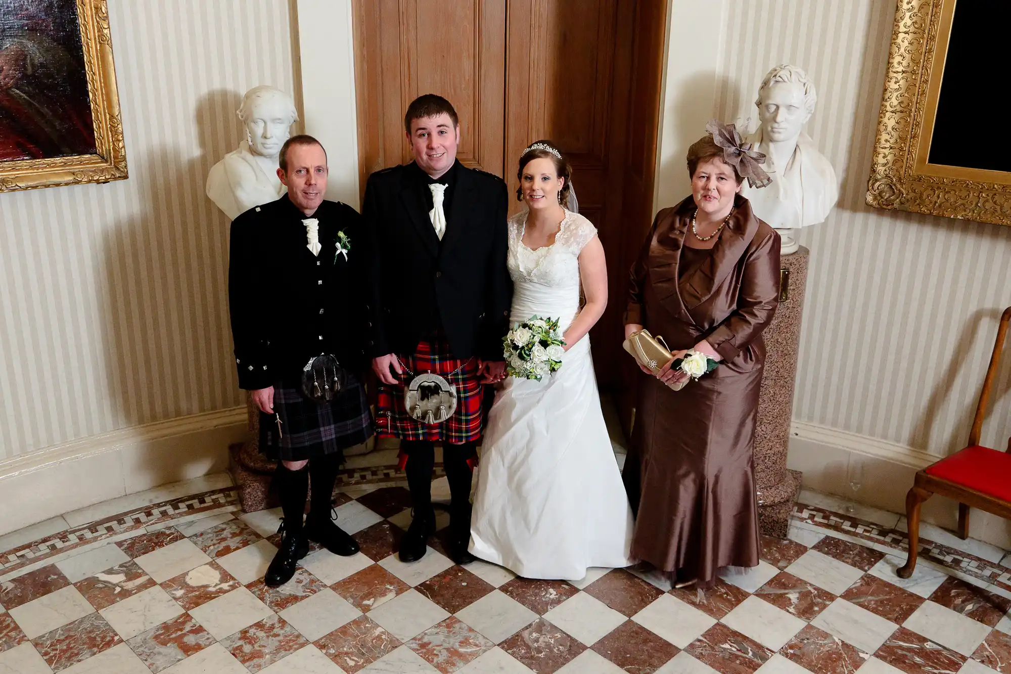 A bride and groom pose with two guests in formal attire, including kilts, in a room with classical busts and a checkered floor.