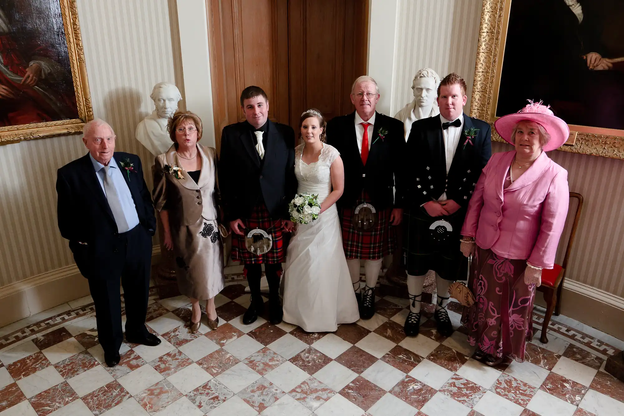A formal family portrait at a wedding with eight people, some wearing kilts and others in traditional dresses, standing in a room with classical decor.