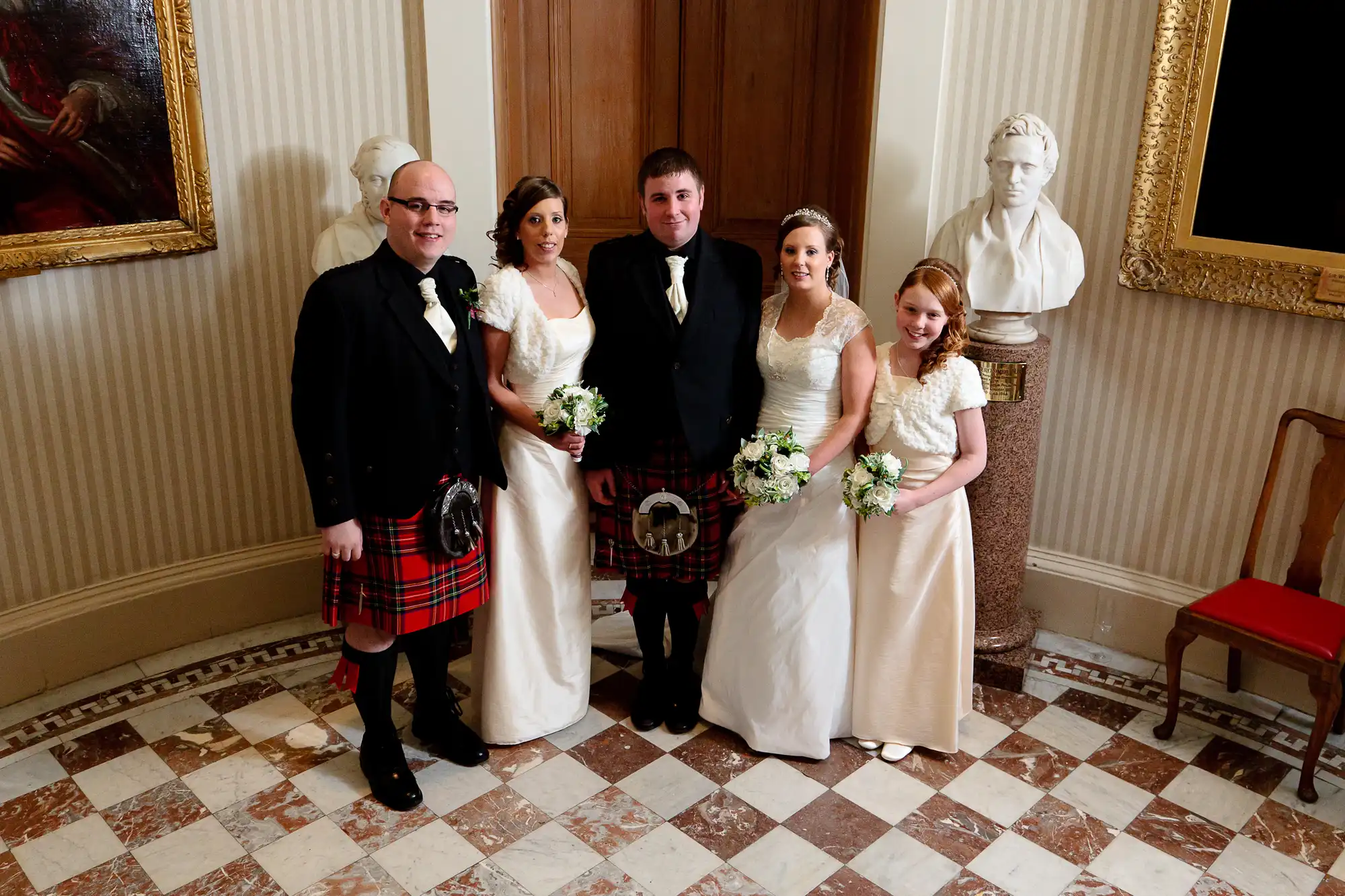 Five individuals in formal wedding attire, two men in kilts, pose together in an elegant room with classical bust sculptures and paintings.