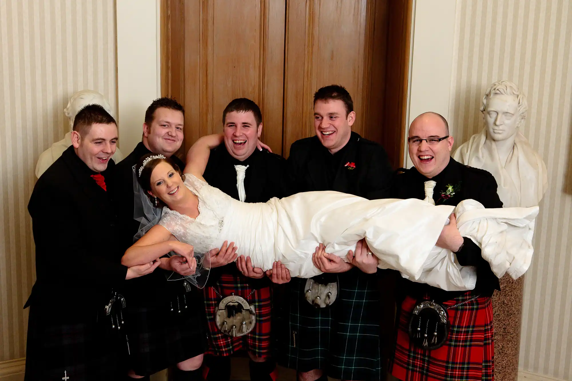 A bride in a white dress is playfully held horizontally by five men in kilts and formal jackets, all smiling in a room with wooden paneling and a bust sculpture.