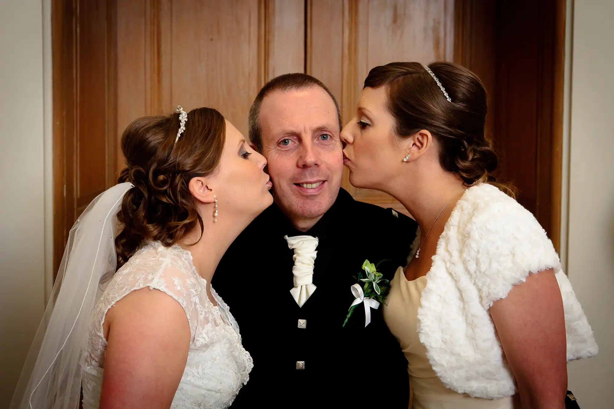 Two brides kissing a smiling groom on his cheeks in a wedding setting. all three are dressed in formal wedding attire.