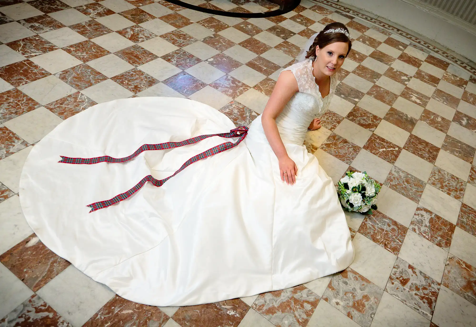 Bride sitting on a tiled floor in a large ballroom, wearing a white gown with an expansive train and holding a bouquet of white flowers.