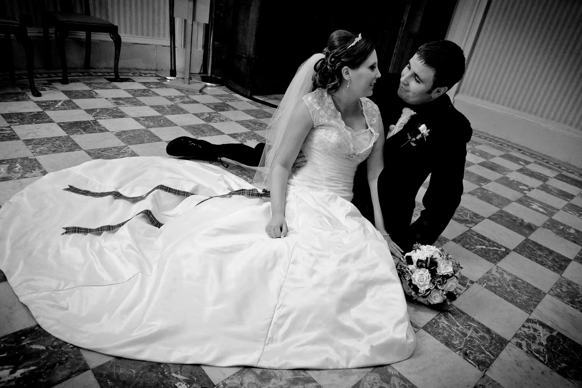 A bride in a white gown and a groom in a suit sitting closely on a checkered floor, gazing into each other's eyes.