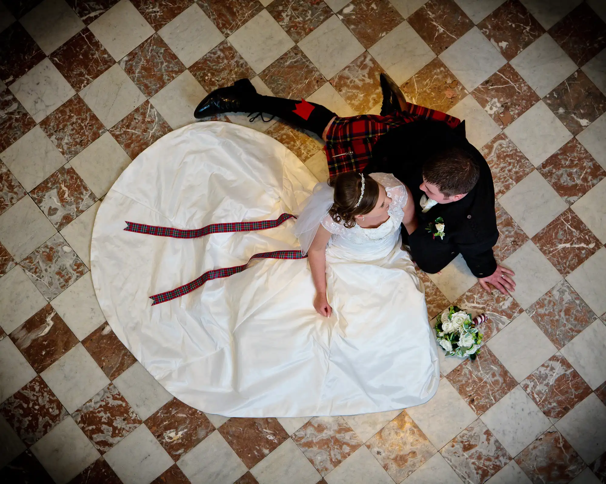 A bride in a white dress and a groom in a kilt lie on a checkered floor, gazing into each other’s eyes, with a bouquet nearby.