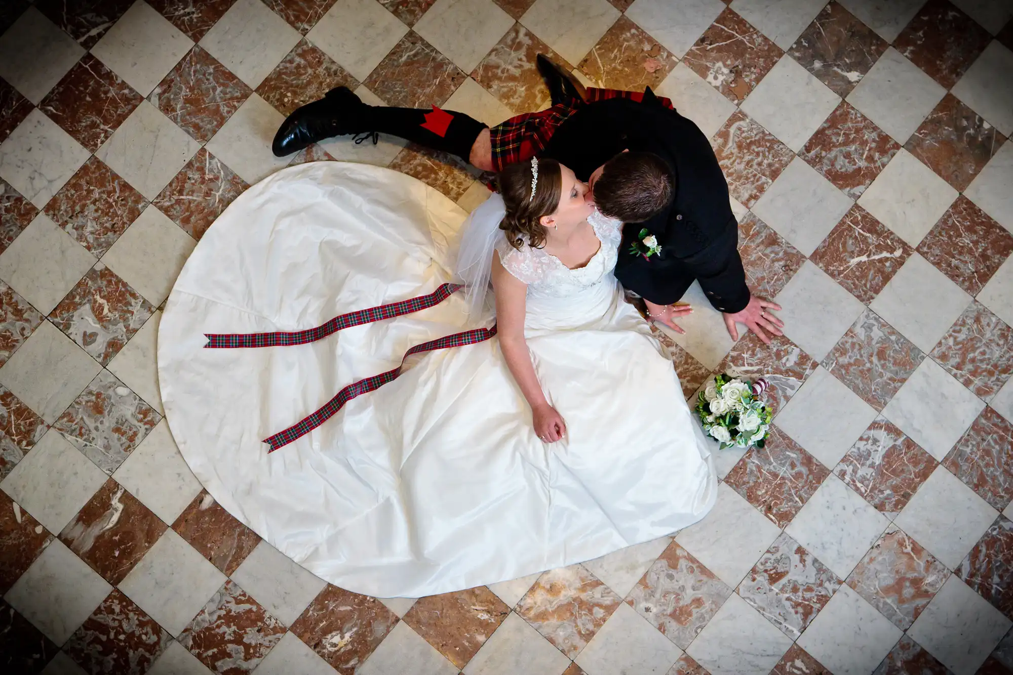A bride and groom in wedding attire sharing a kiss on a checkered floor; the groom wears a kilt and the bride's dress has a long train.