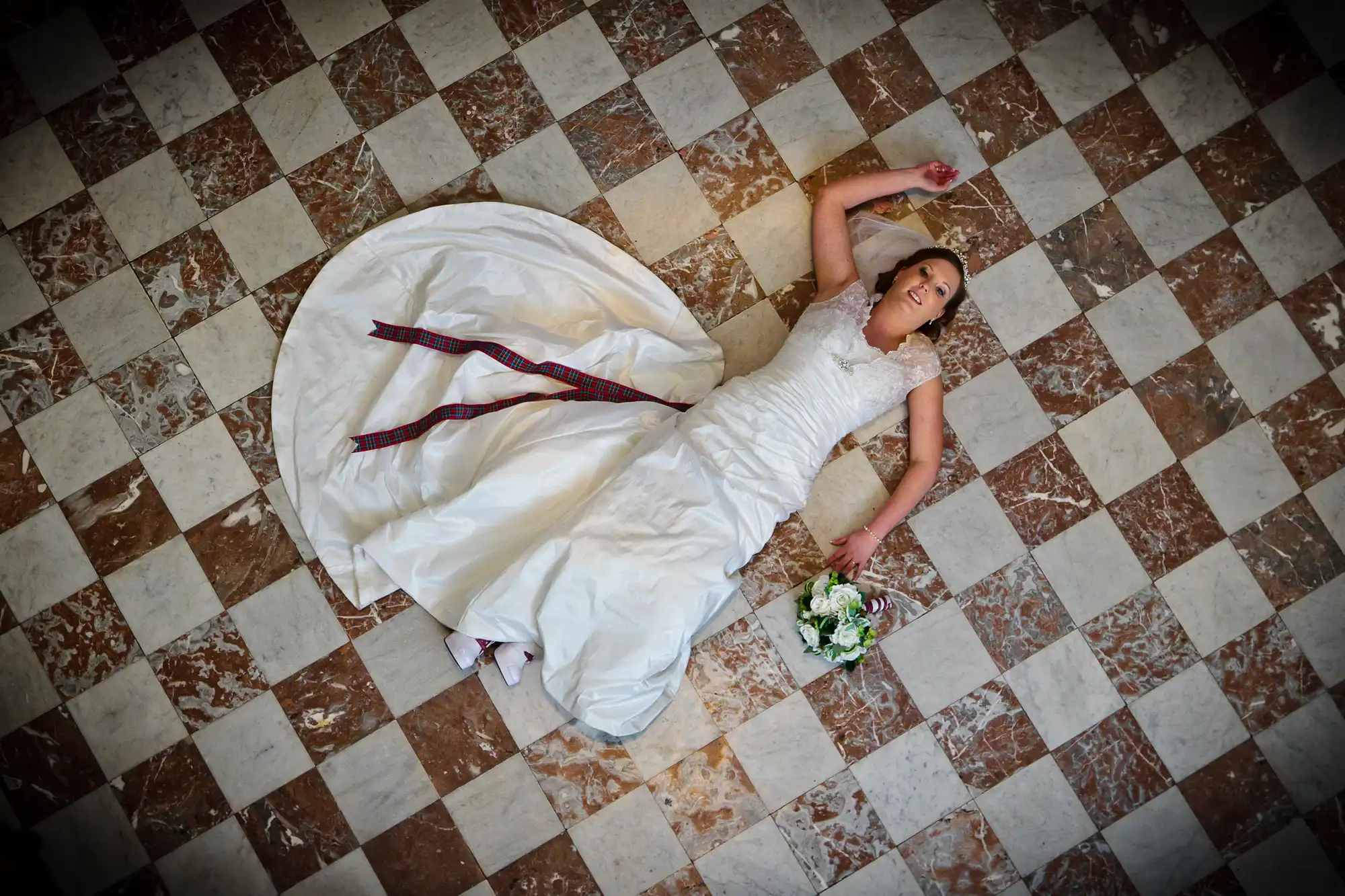 A bride in a white gown with a red ribbon lies on a checkered floor, holding a bouquet, looking upwards.