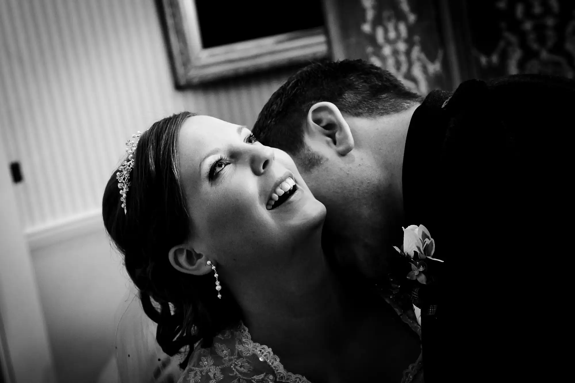 Black and white photo of a bride smiling at her groom as he whispers in her ear, both dressed in wedding attire.