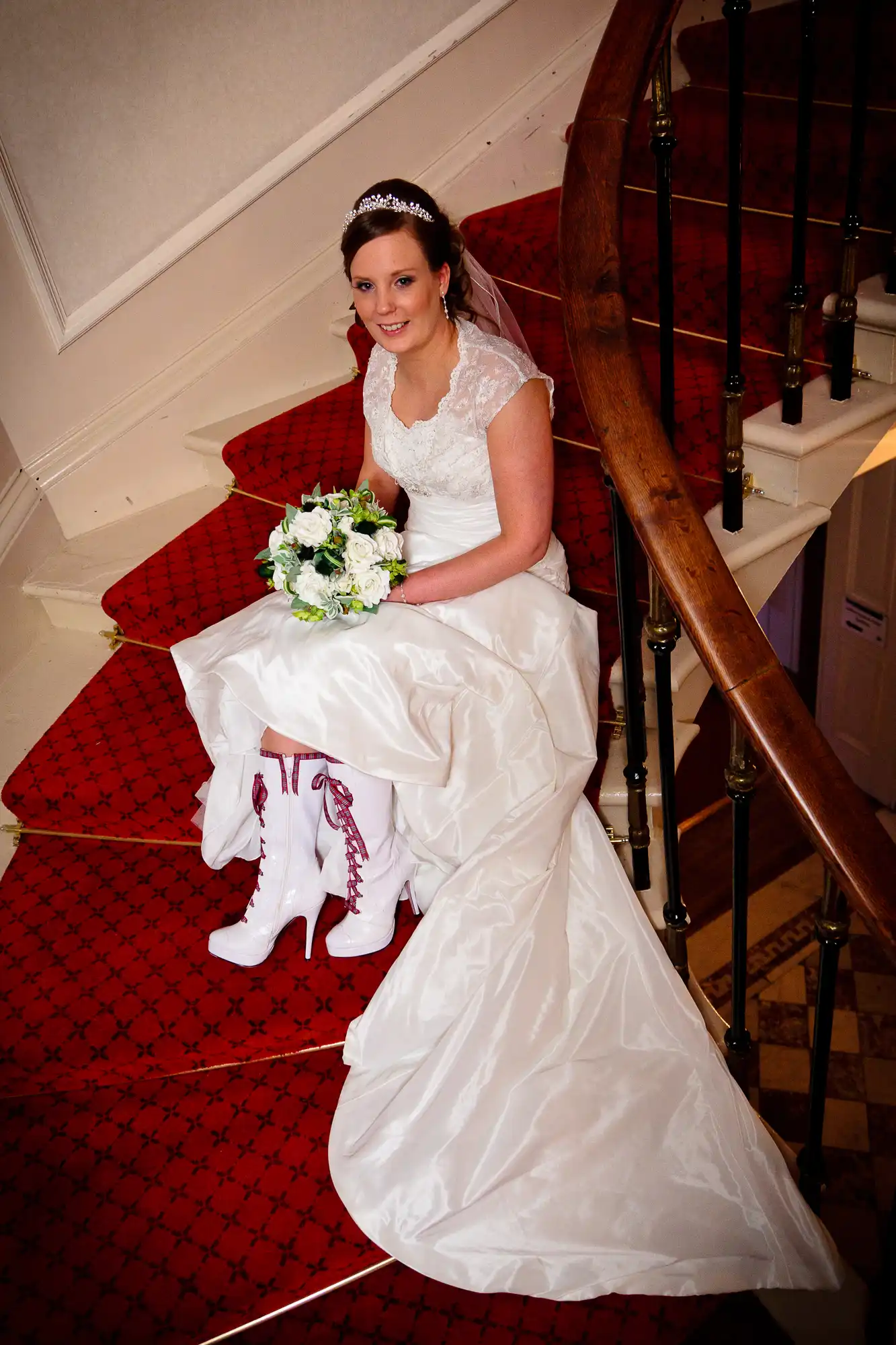 A bride sitting on a staircase in a white dress, holding a bouquet, with a patterned red carpet.