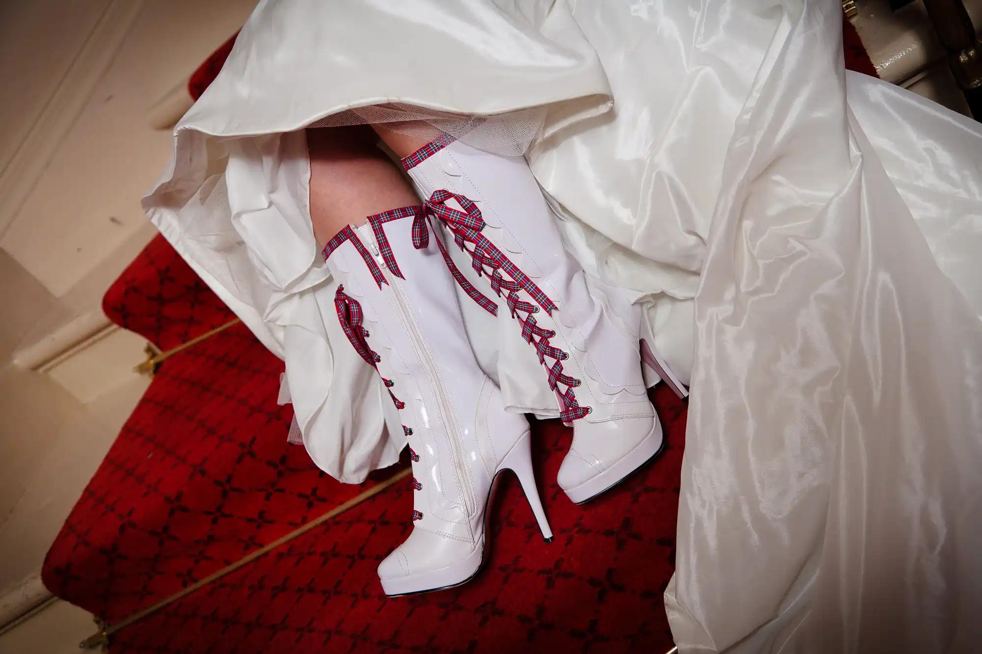 A bride wearing white boots with red ribbon lacing under her lifted wedding dress on a red carpeted staircase.