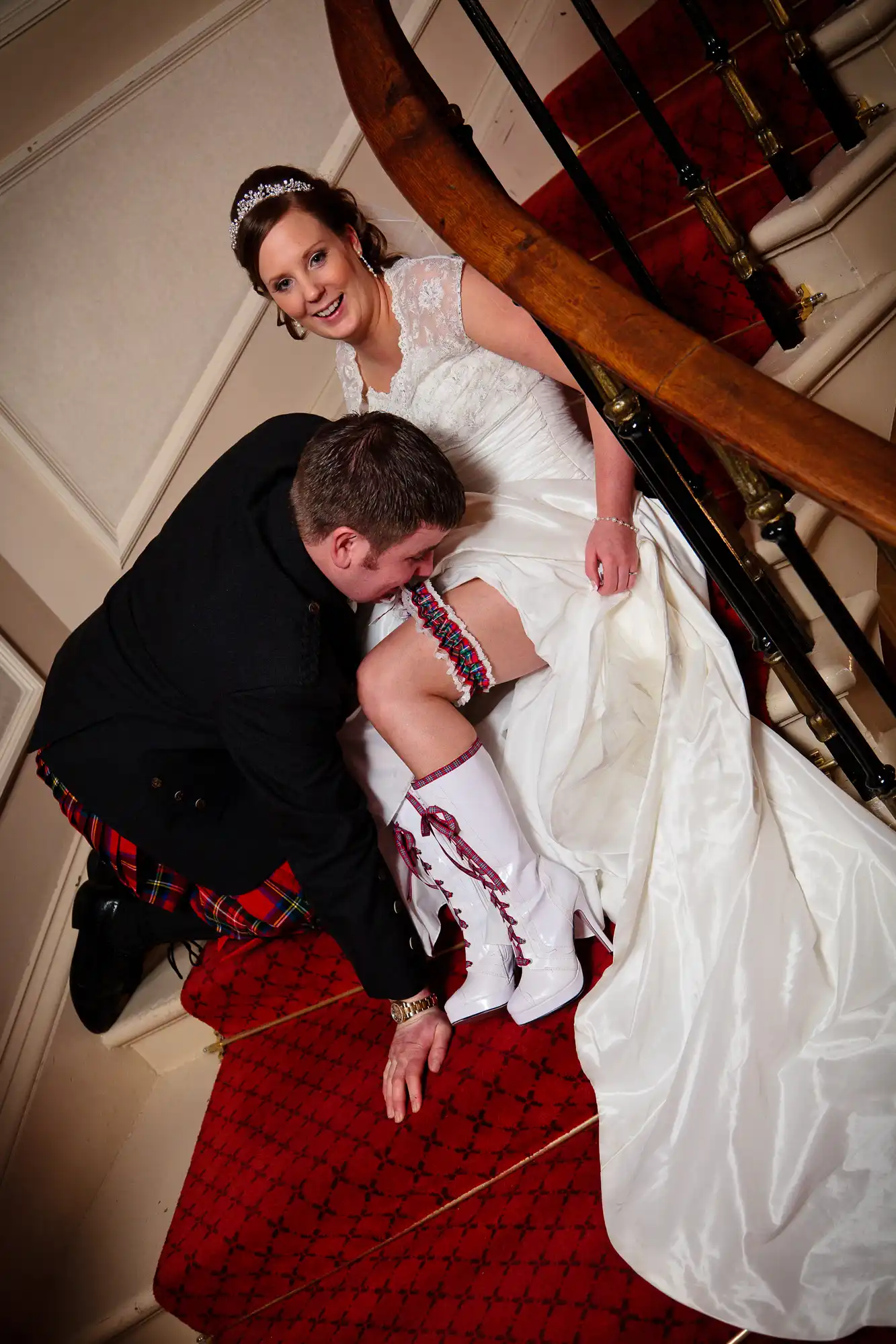 A bride in a white dress sitting on a staircase, smiling as the groom in a kilt adjusts her white garter.