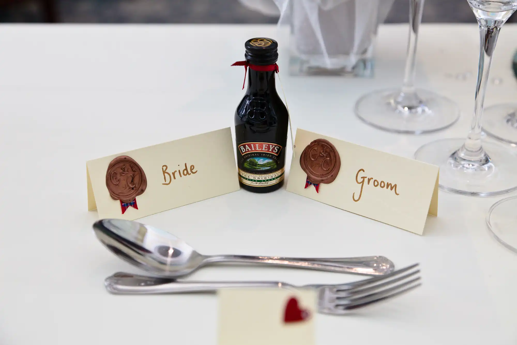Place cards labeled "bride" and "groom" with wax seals flank a miniature baileys bottle on a dining table, complete with silverware and a heart-shaped decoration.