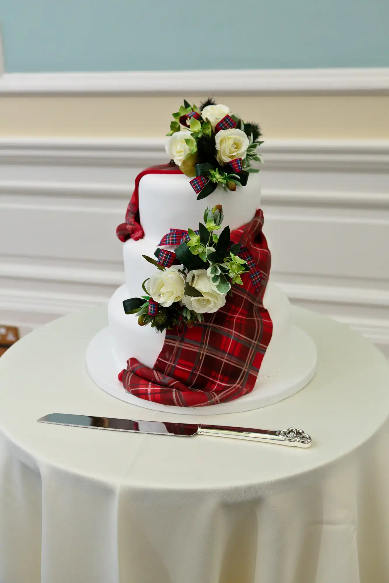 A three-tier wedding cake adorned with white icing, red tartan ribbons, and a floral arrangement, on a table beside a cake knife.
