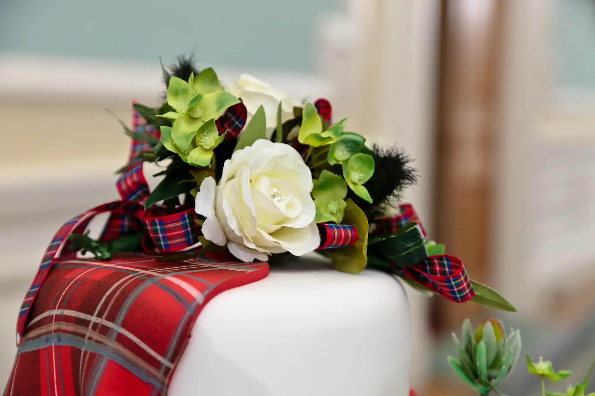 A bouquet of white flowers and greenery wrapped with a tartan ribbon, placed atop a red plaid gift box.
