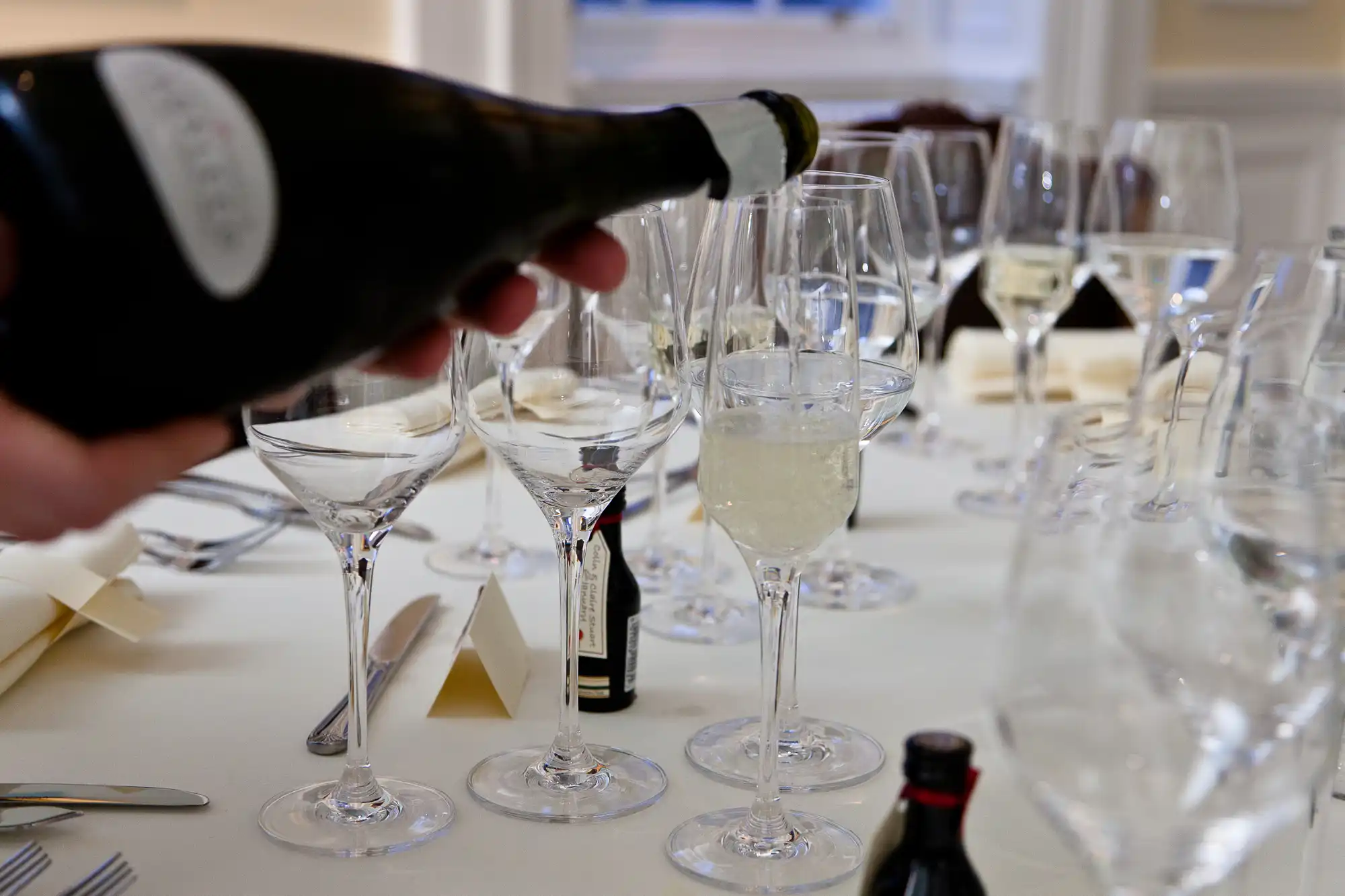A person pouring champagne into flute glasses at a table set with multiple empty glasses and folded napkins.