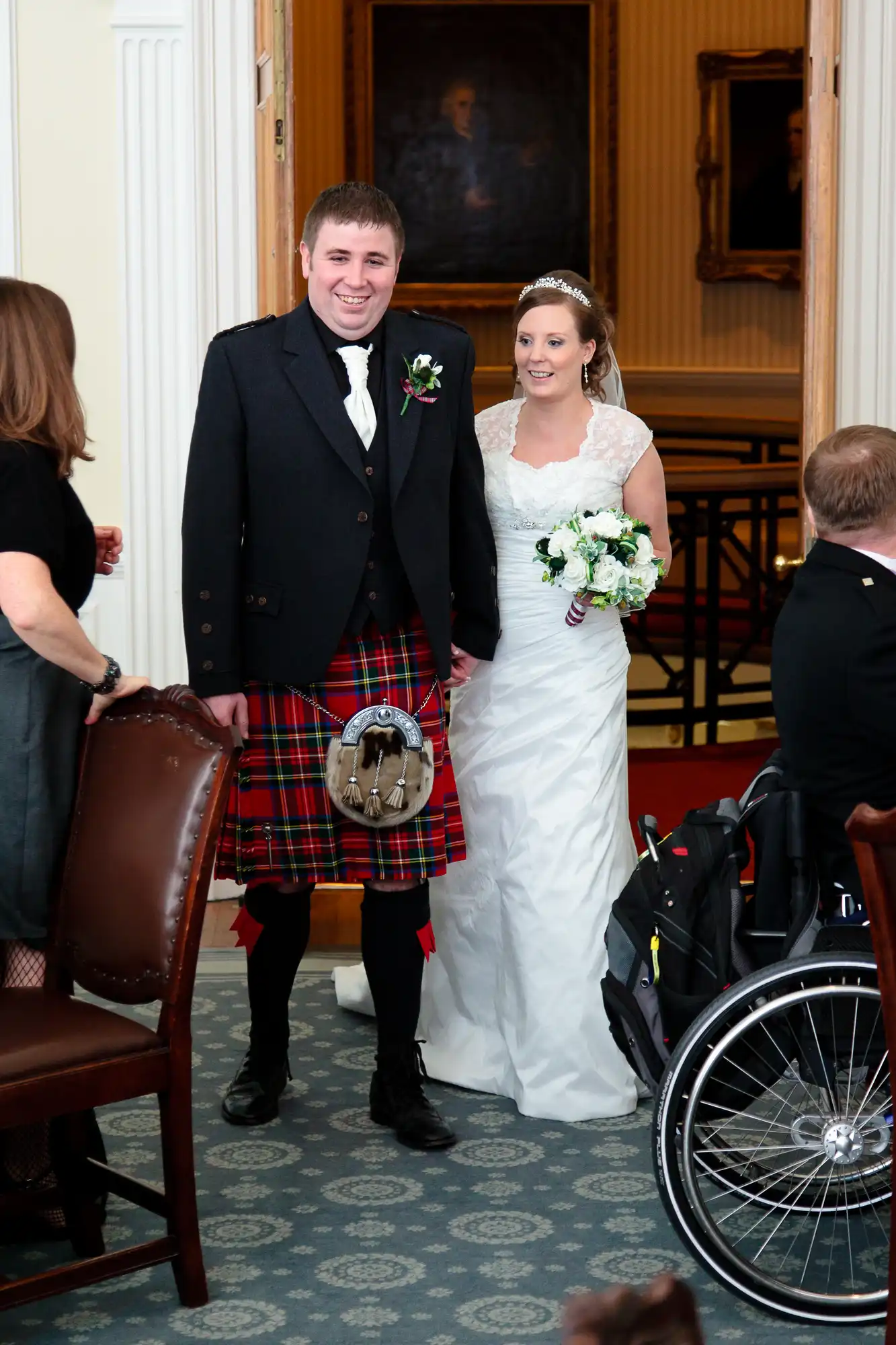 A bride and groom smiling as they walk together, the groom in a traditional scottish kilt and the bride in a white mermaid-style gown, holding a bouquet.