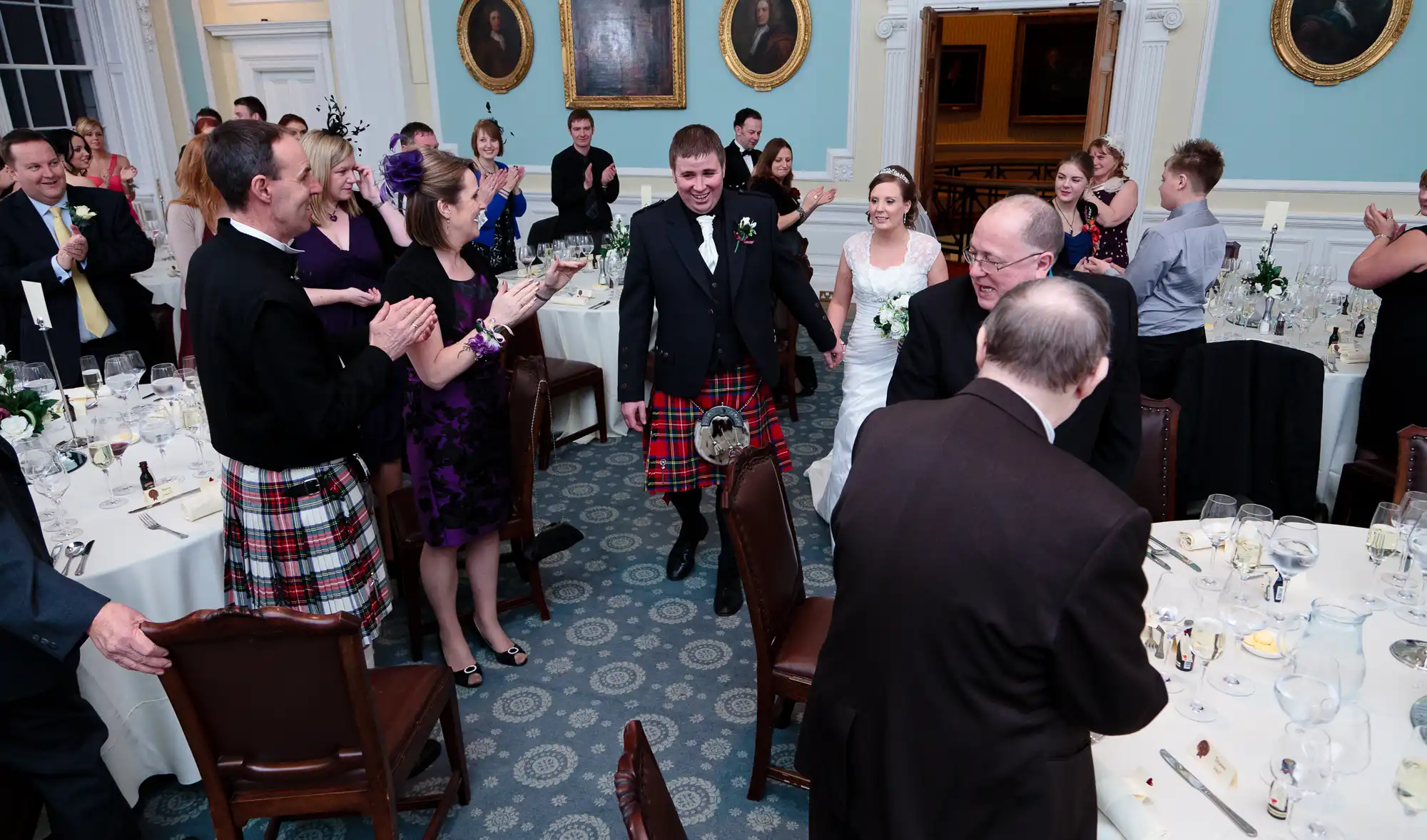 A bride and groom, the groom in a kilt, enter a banquet room to applause from guests seated at decorated tables.