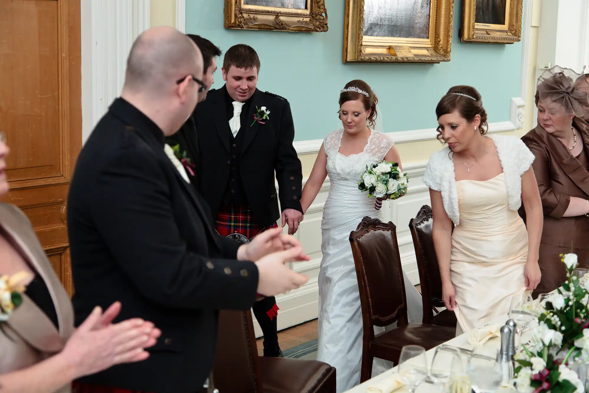 A bride and groom, followed by guests, walk through a reception room as a man gestures towards an empty chair.