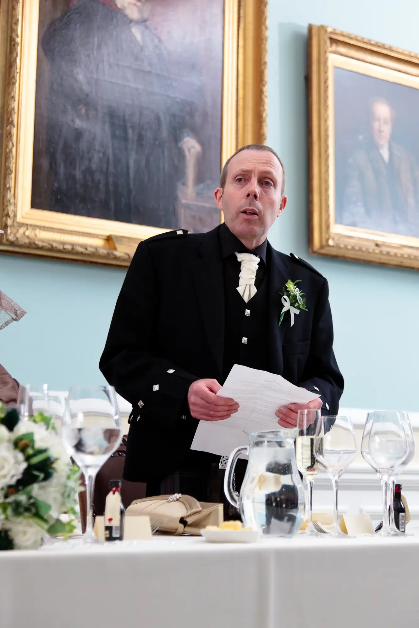 A man in a formal black suit and boutonniere giving a speech at a wedding reception, holding a paper, with paintings in the background.