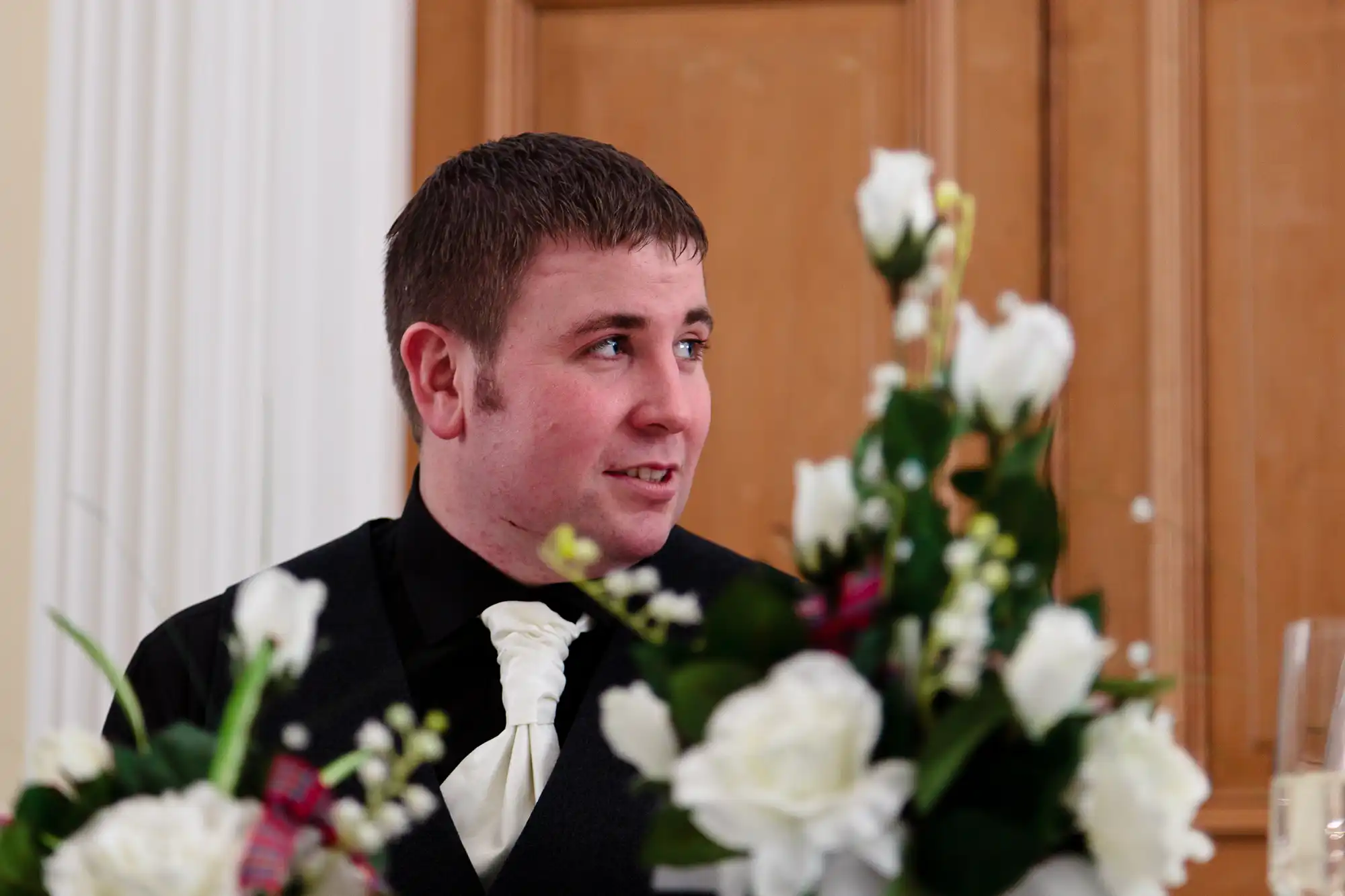 Man in a black tuxedo sitting at a table, smiling slightly and looking to the side, with white roses in the foreground.