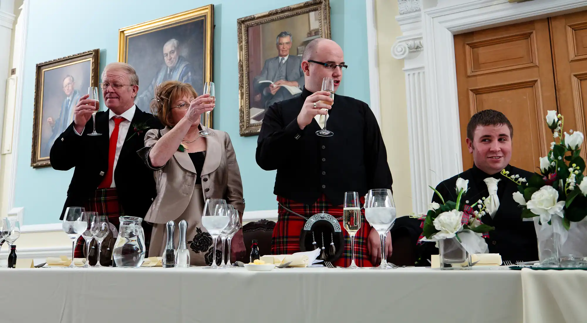Four people at a formal event, two in kilts, raising glasses in a toast; portraits on the wall behind them.