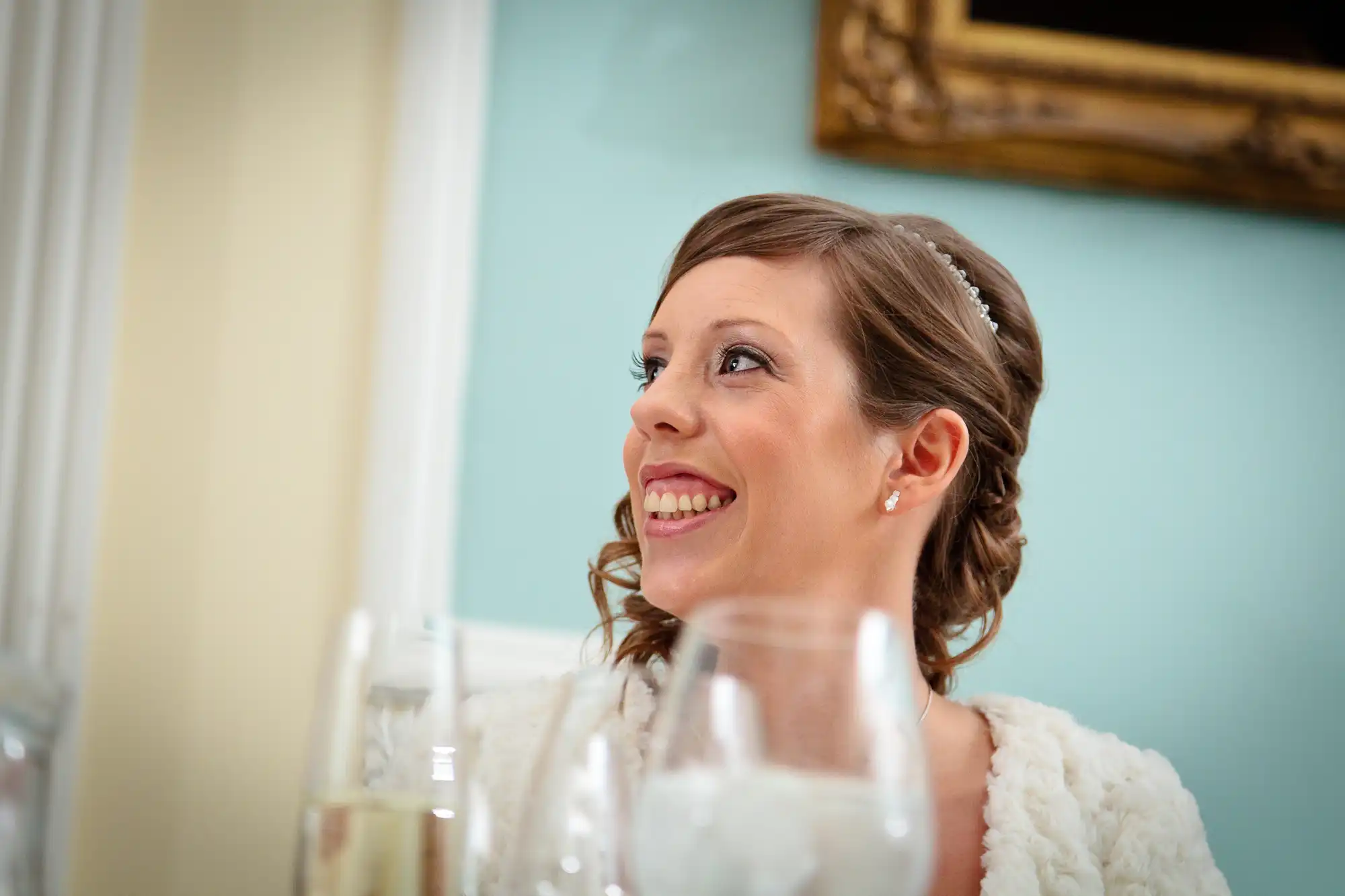 A smiling bride, wearing a tiara and white dress, looks away thoughtfully in a room with light blue walls and elegant decor.