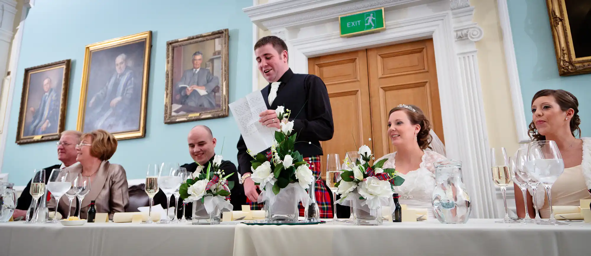 A man standing and giving a speech at a wedding reception table, with guests seated around listening, in a room with portraits on the walls.