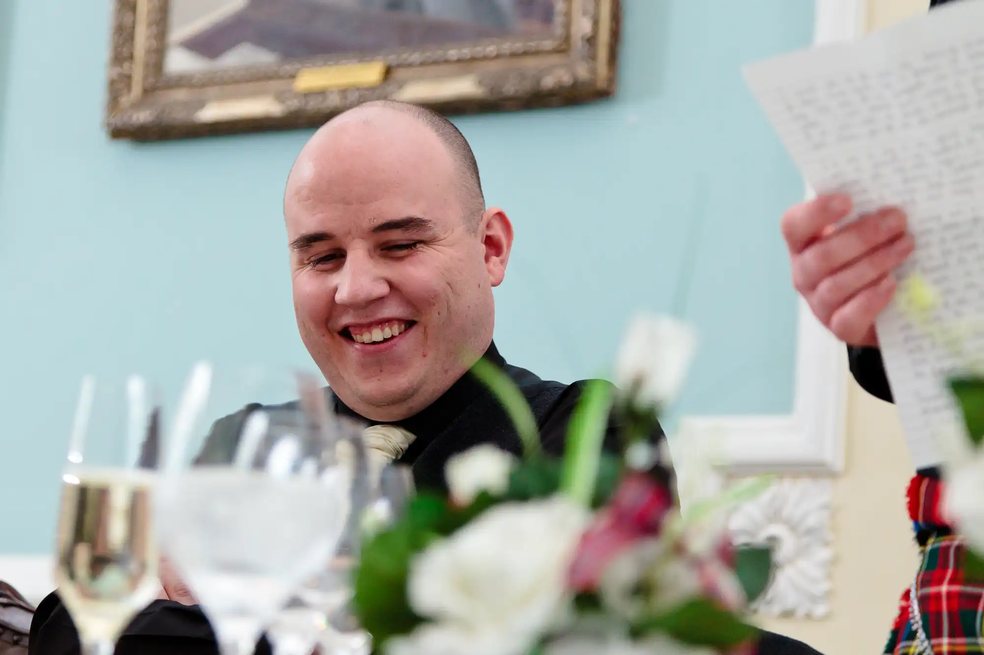A bald man in a formal outfit smiles while seated at a table with flowers and wine glasses, with another person holding a paper nearby.