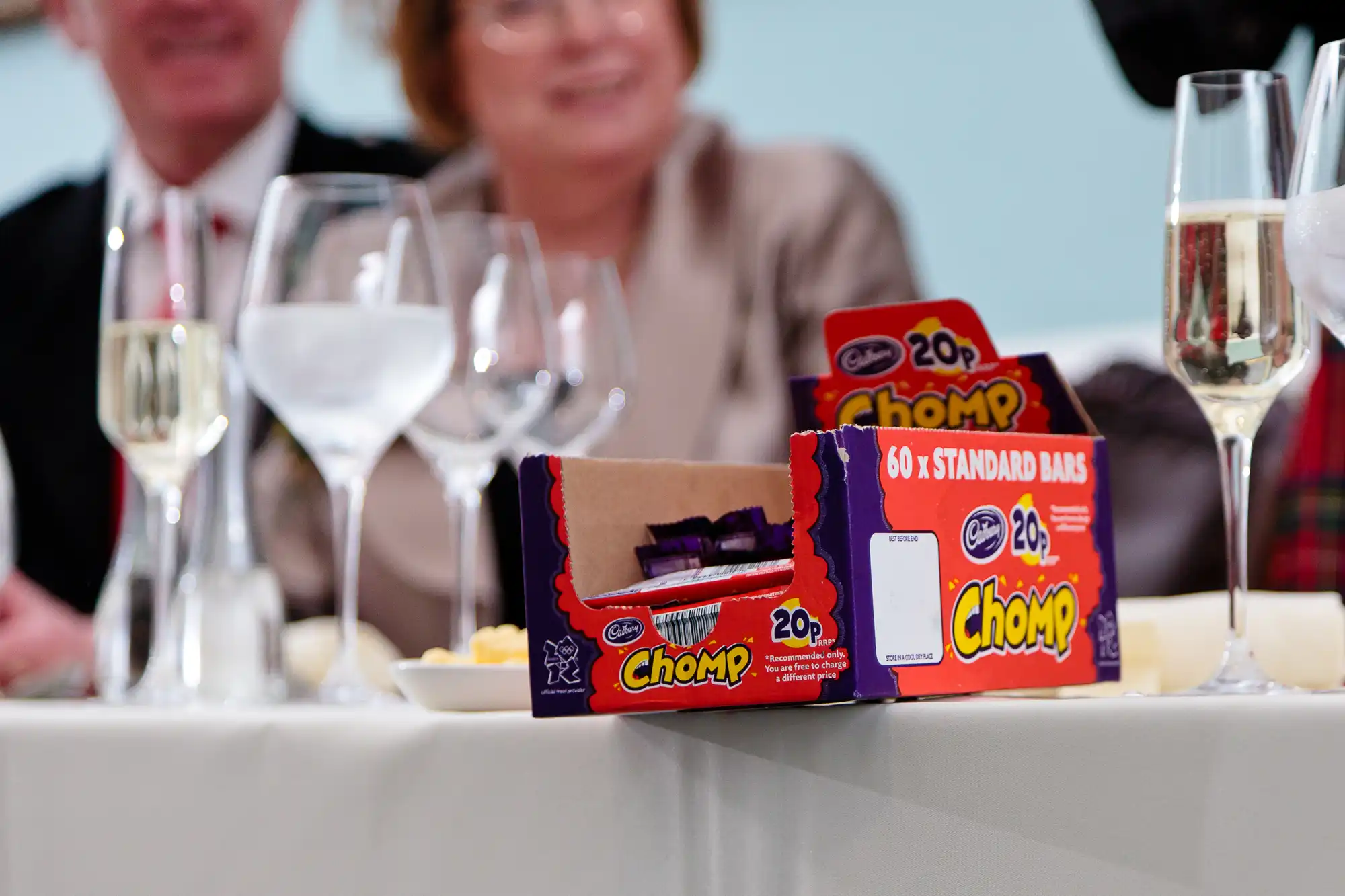 A box of cadbury chomp chocolate bars on a table at an event, with blurred guests and wine glasses in the background.