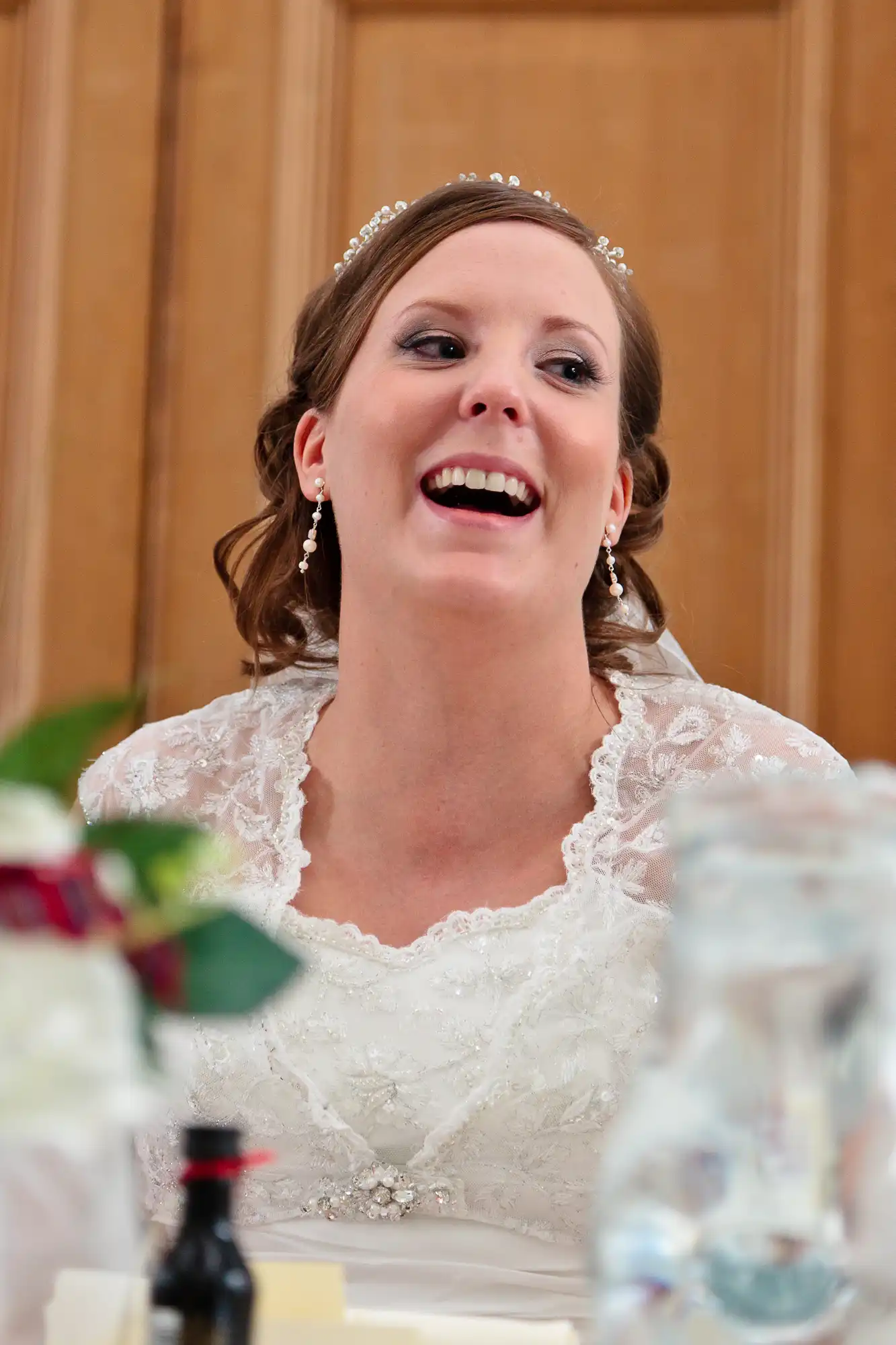 A bride in a lace wedding dress, smiling broadly at a table during a reception, adorned with a tiara and earrings.