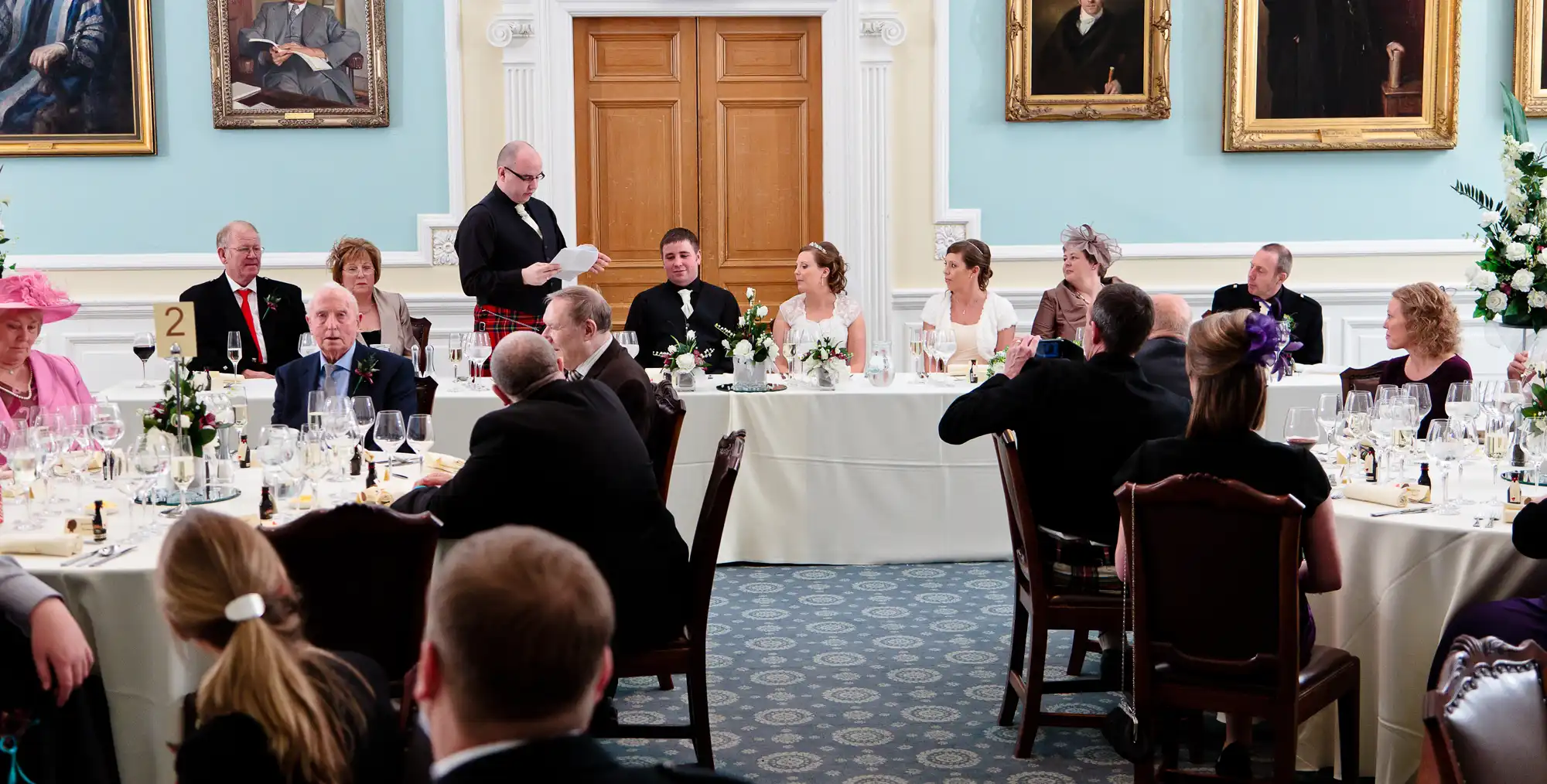 Wedding guests seated at a formal dinner, watching a man giving a speech at the head table, in a room adorned with portraits.