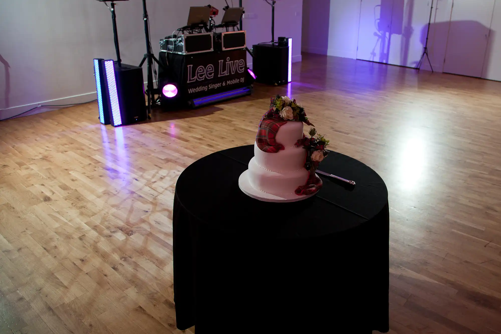A wedding cake decorated with tartan ribbon and flowers on a table, with a dj booth titled "lee live" in the background of a dimly lit room.