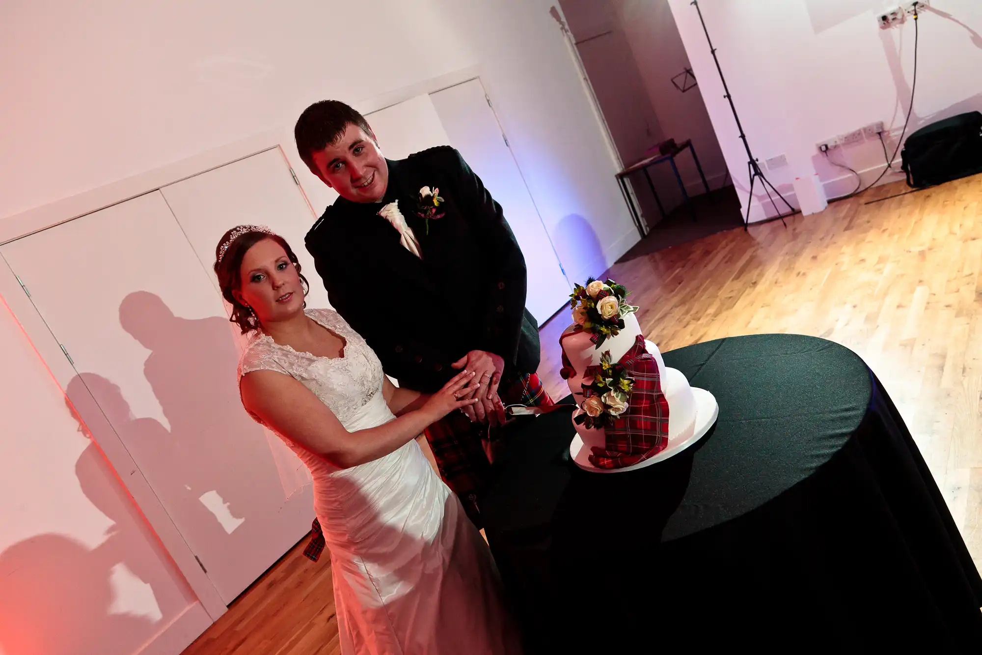 A bride and groom cutting a wedding cake in a banquet hall, both smiling at the camera.