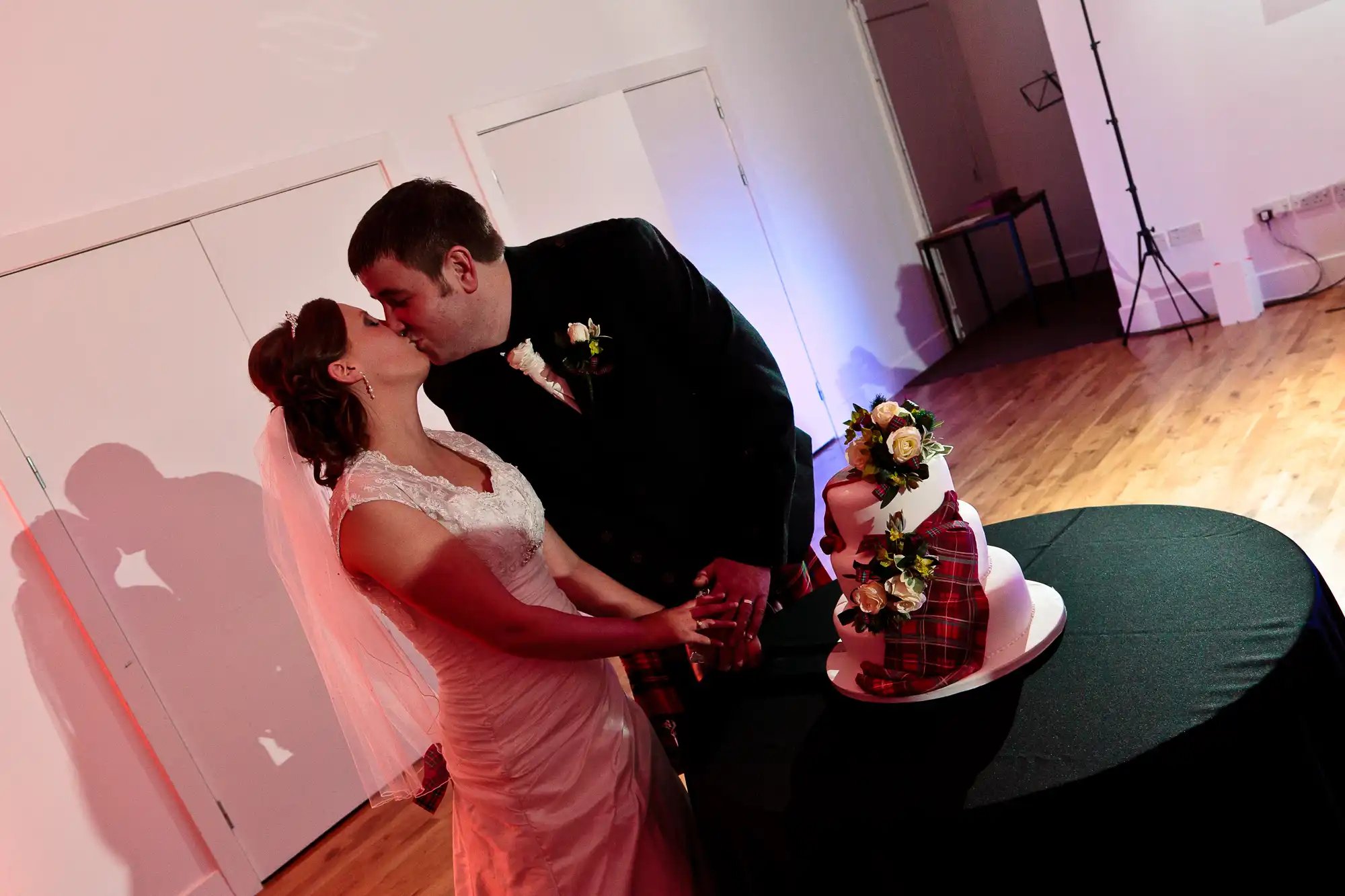 Bride and groom kissing beside their wedding cake in a dimly lit room, with their shadows cast on a white wall.
