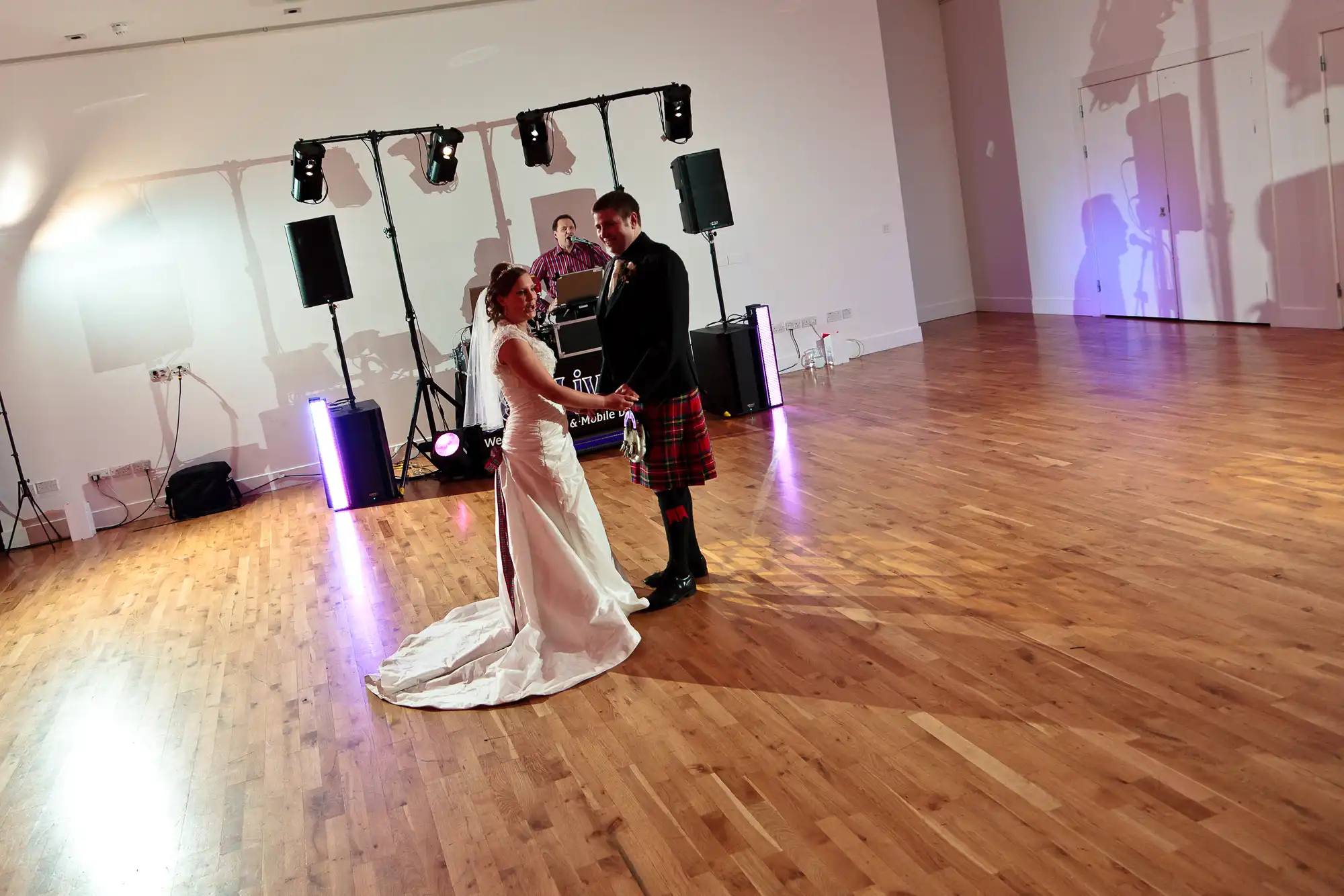 A bride in a white dress and a groom in a traditional kilt dance together in a well-lit hall with a dj setup in the background.