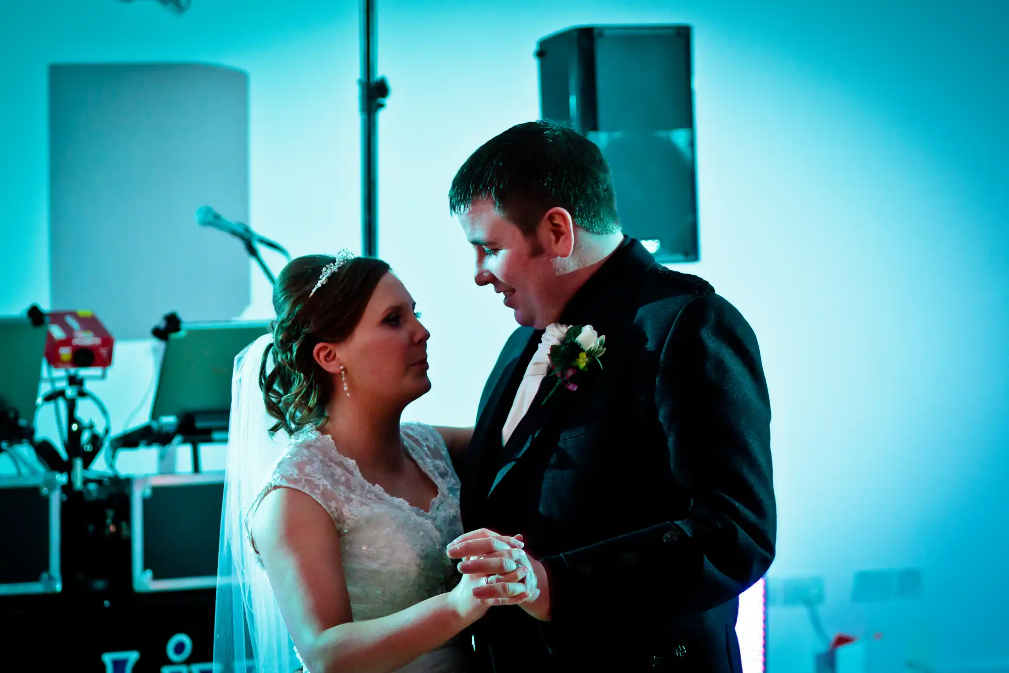 A bride and groom share an intimate dance under blue lighting at their wedding reception, with speakers and dj equipment in the background.