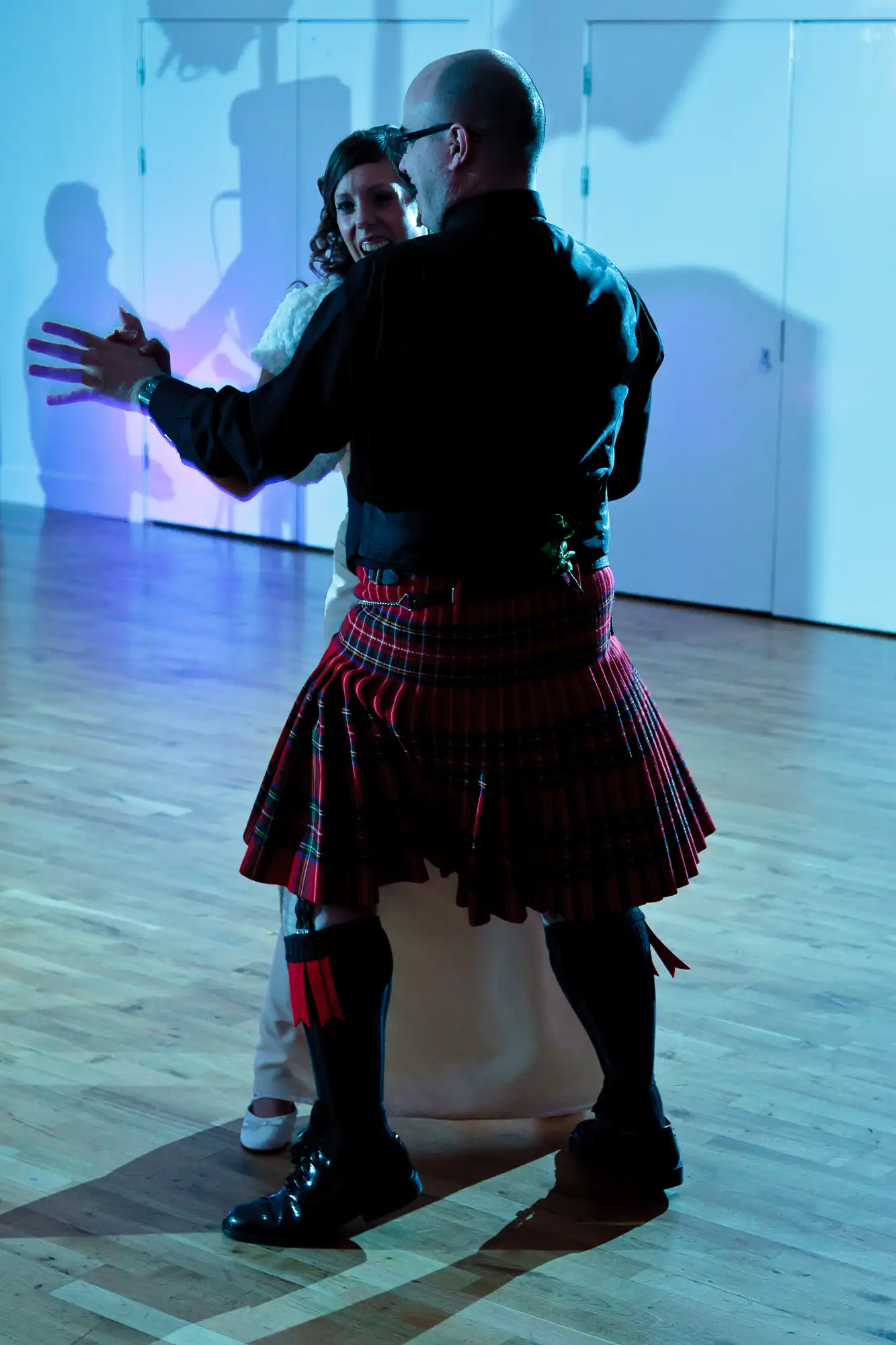 A man in a kilt and a woman dancing closely in a room with blue lighting and reflective walls.