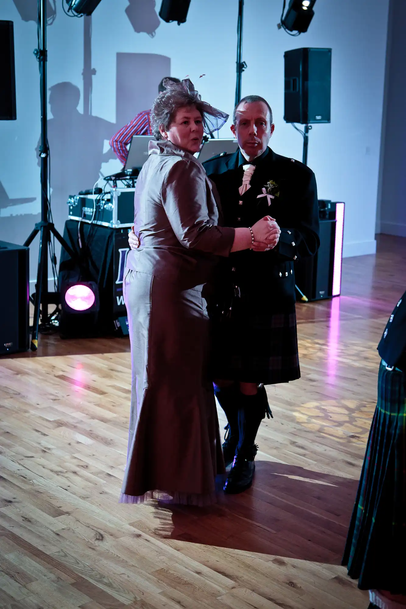 A woman in a formal dress dancing with a man wearing a kilt and formal attire at a wedding reception, with a band setup in the background.