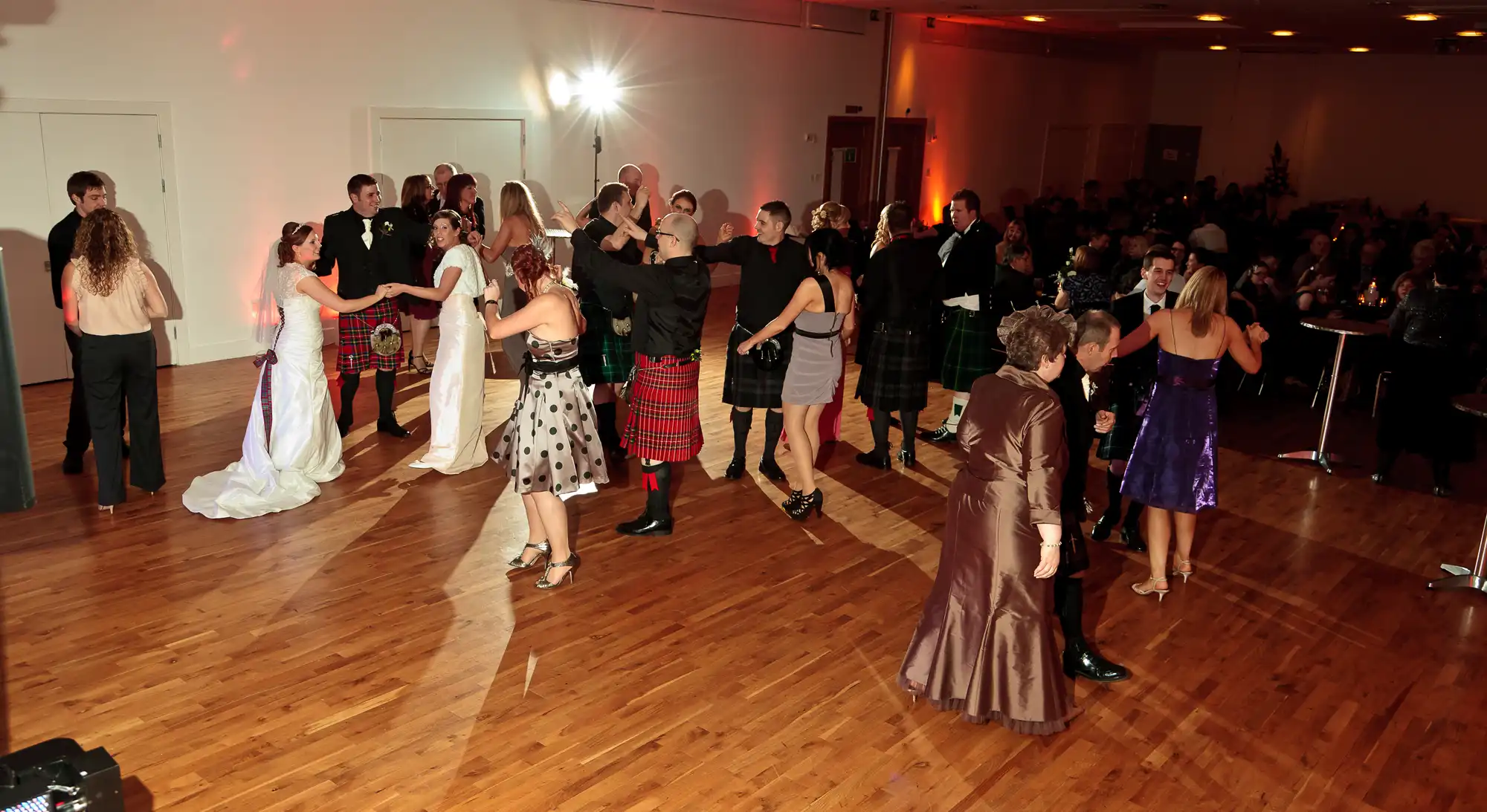 People in formal attire, some wearing kilts, dancing and socializing at a lively indoor gathering.