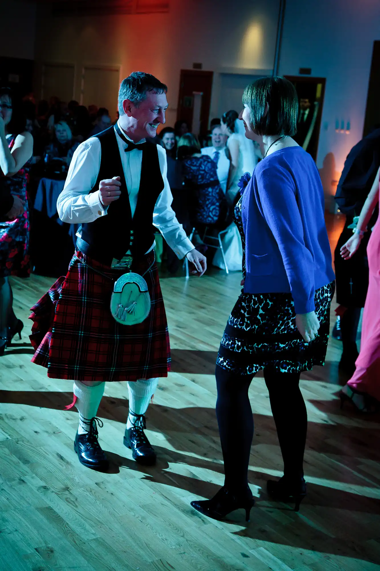 A man in a kilt dances with a woman in a black dress at a lively event with people in the background.