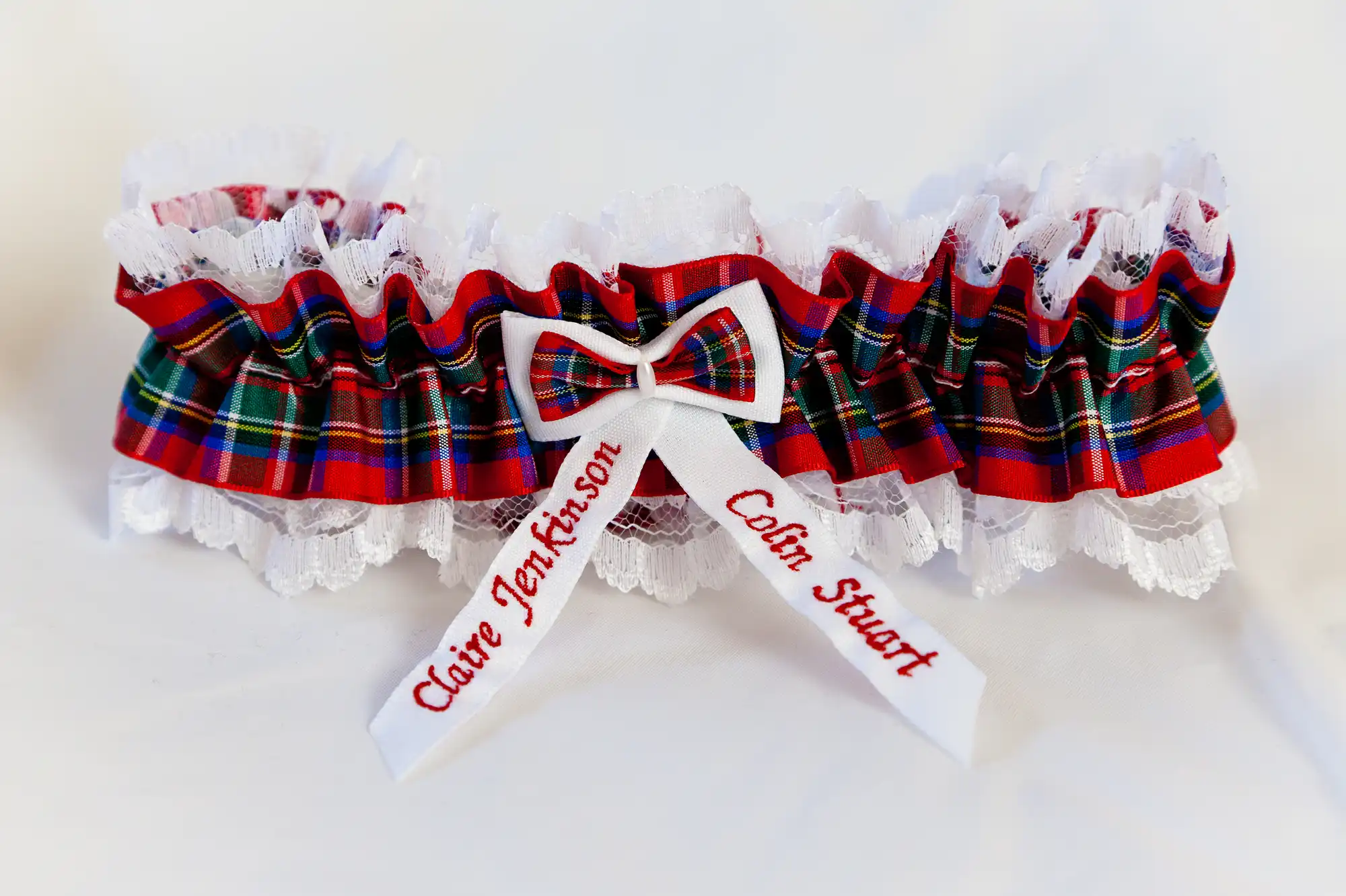 A tartan bridal garter with lace trim, featuring a bow and embroidered with the names "claire jefferson" and "colin stuart.