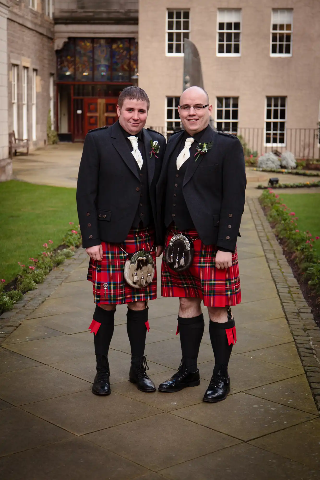 Two men in traditional scottish attire, including kilts and jackets, standing on a paved walkway outside a building with a stained-glass window.