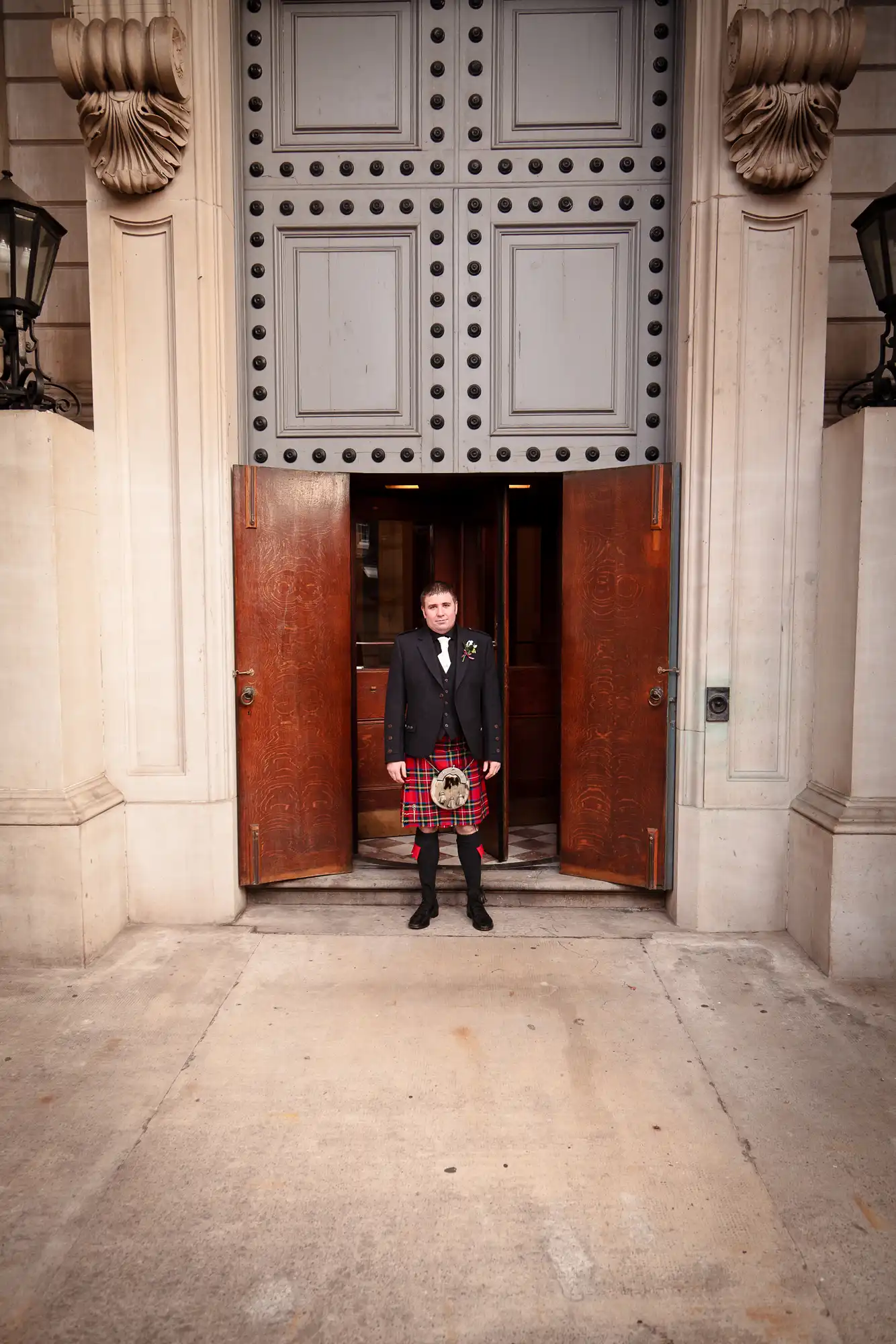 A man in a traditional scottish outfit with a kilt and sporran stands in front of large ornate doors of a building.