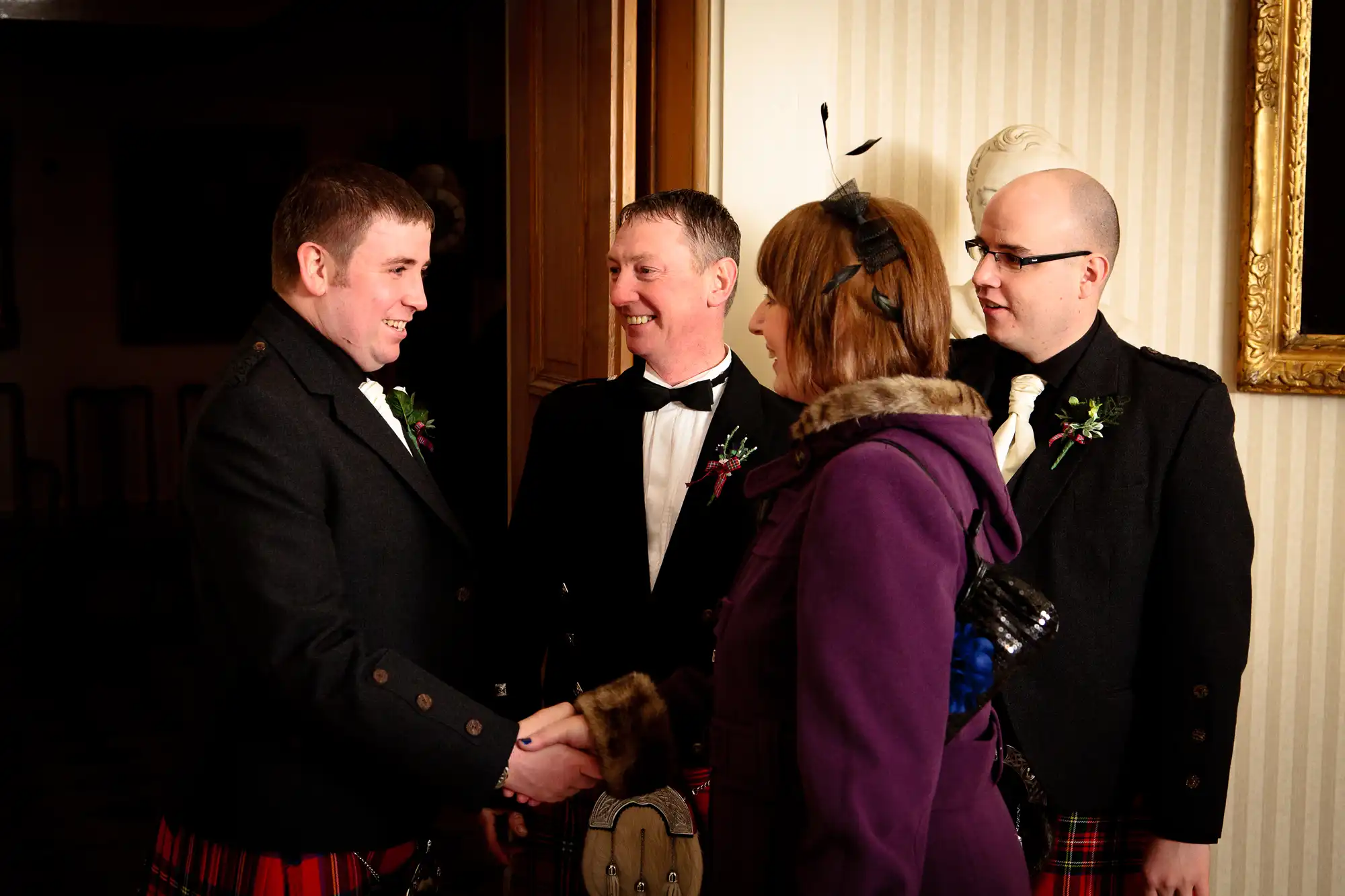 A group of four people, three in kilts, engaging in a friendly handshake and conversation at an indoor event.